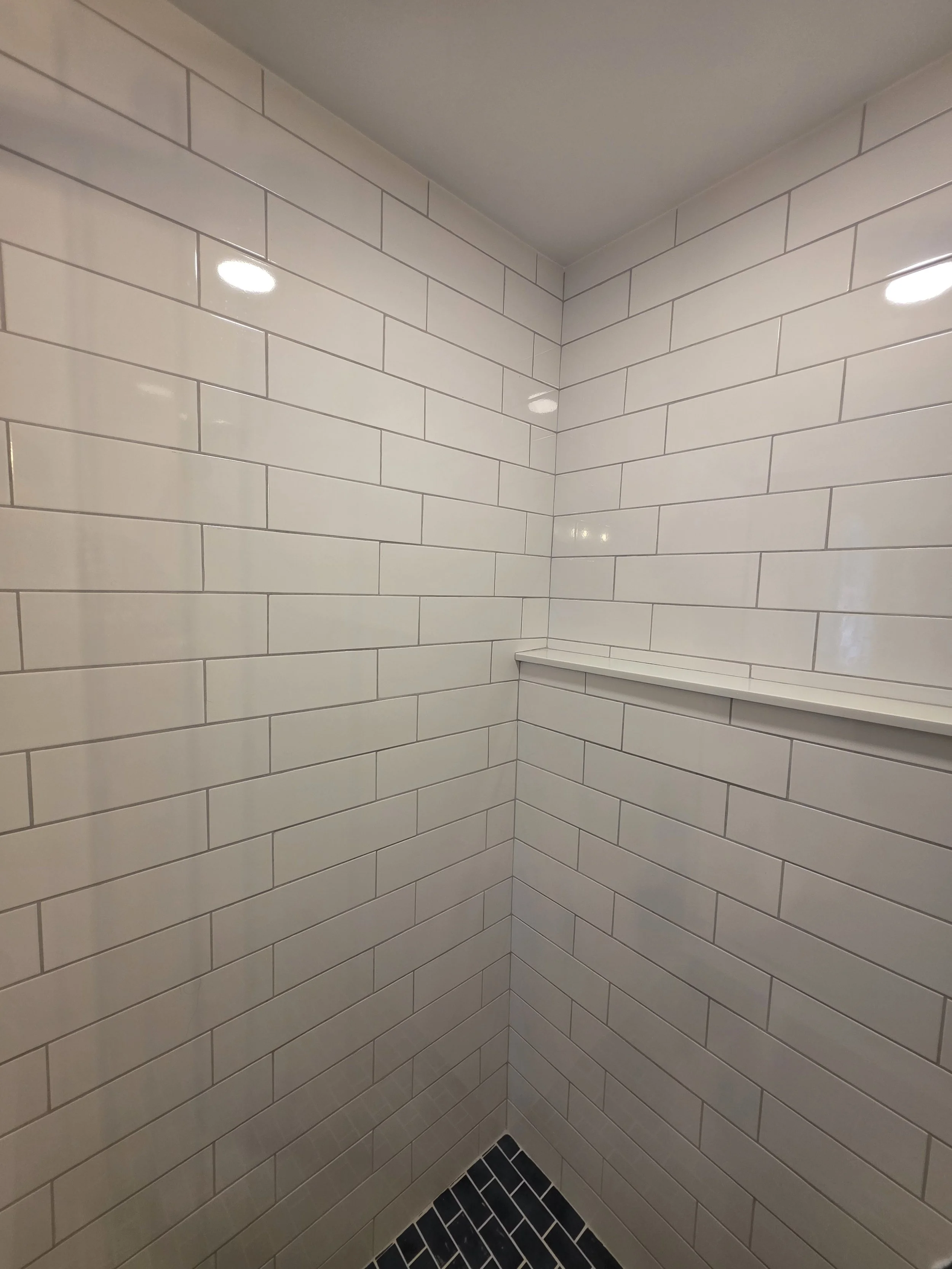 White subway-tile shower corner with a long ledge and dark mosaic floor.