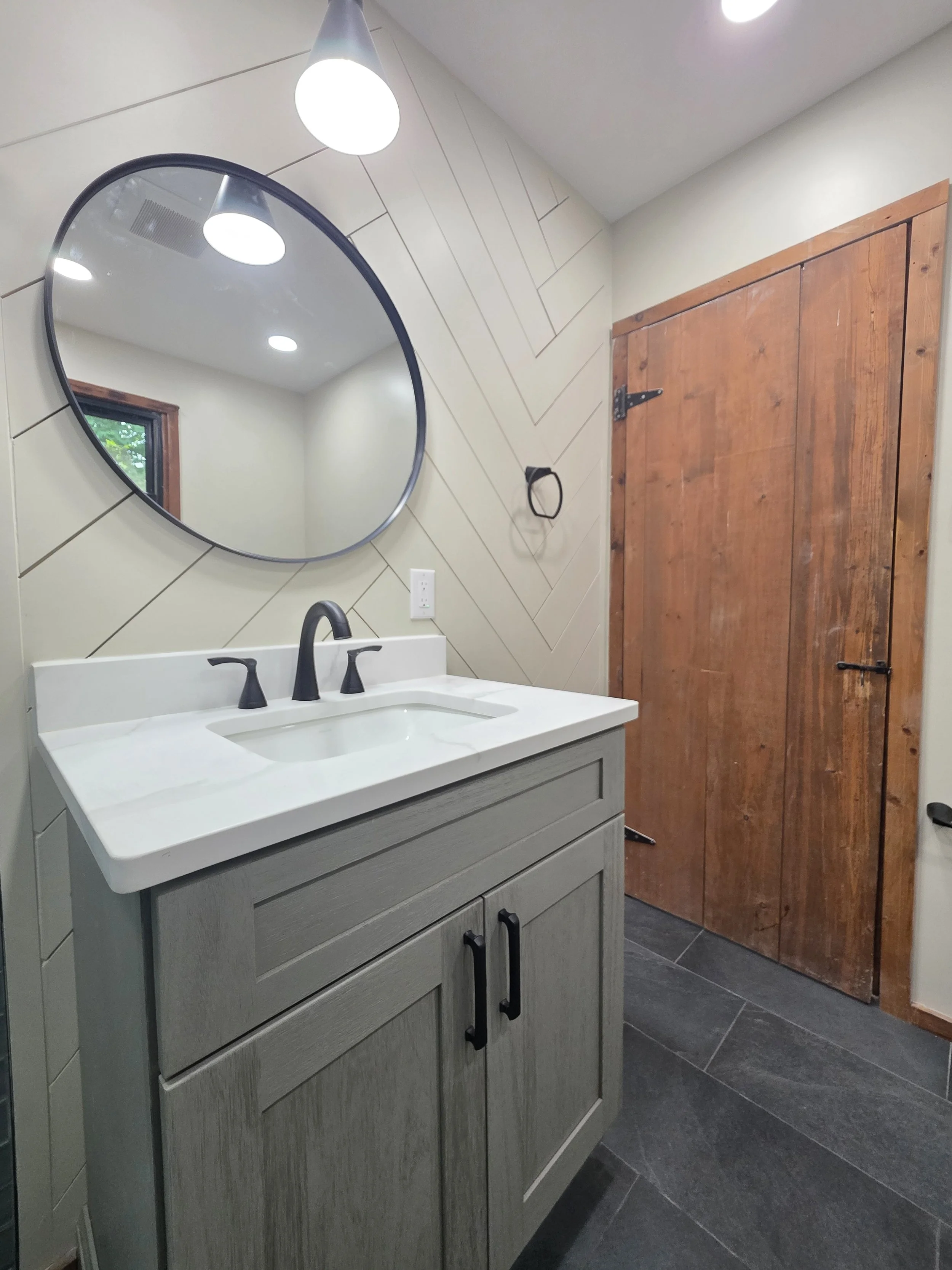 Wood-look vanity with a white countertop, dark faucet and round mirror beside the wood door.