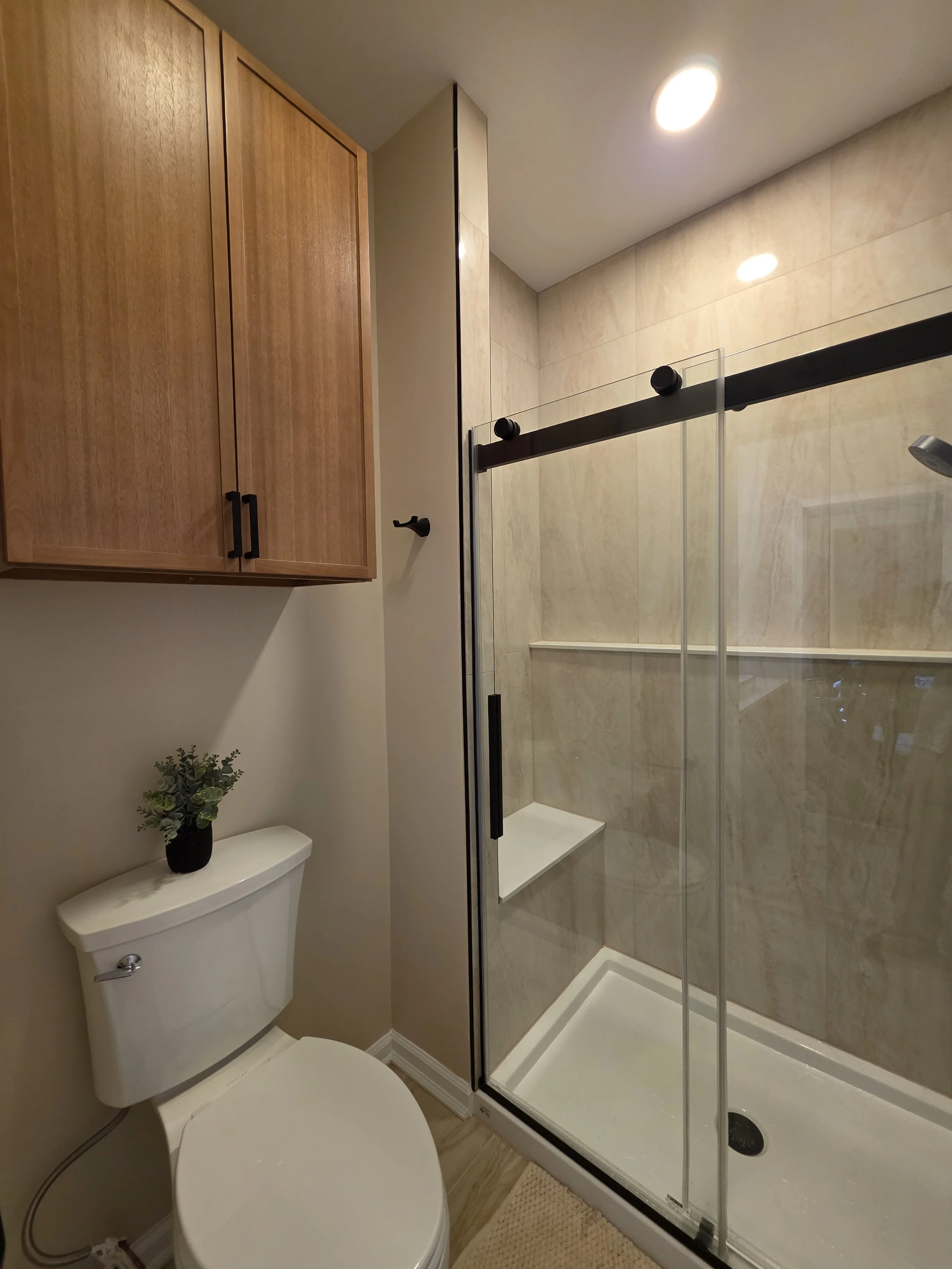Remodeled hallway bathroom with a glass shower, black fixtures and warm wood-tone storage cabinet above the toilet.