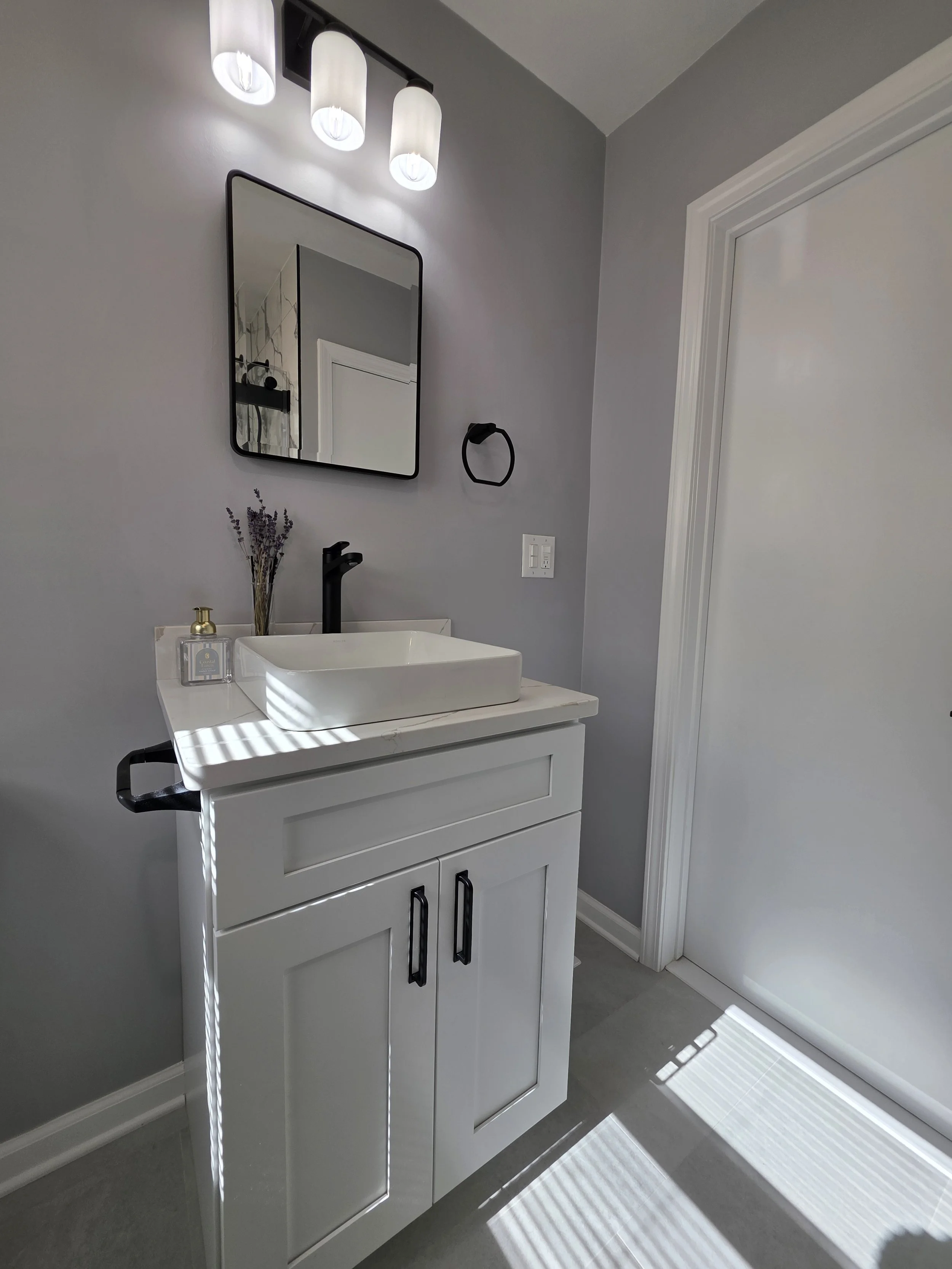 White vanity with a rectangular vessel sink, black hardware and wall mirror beneath three lights.