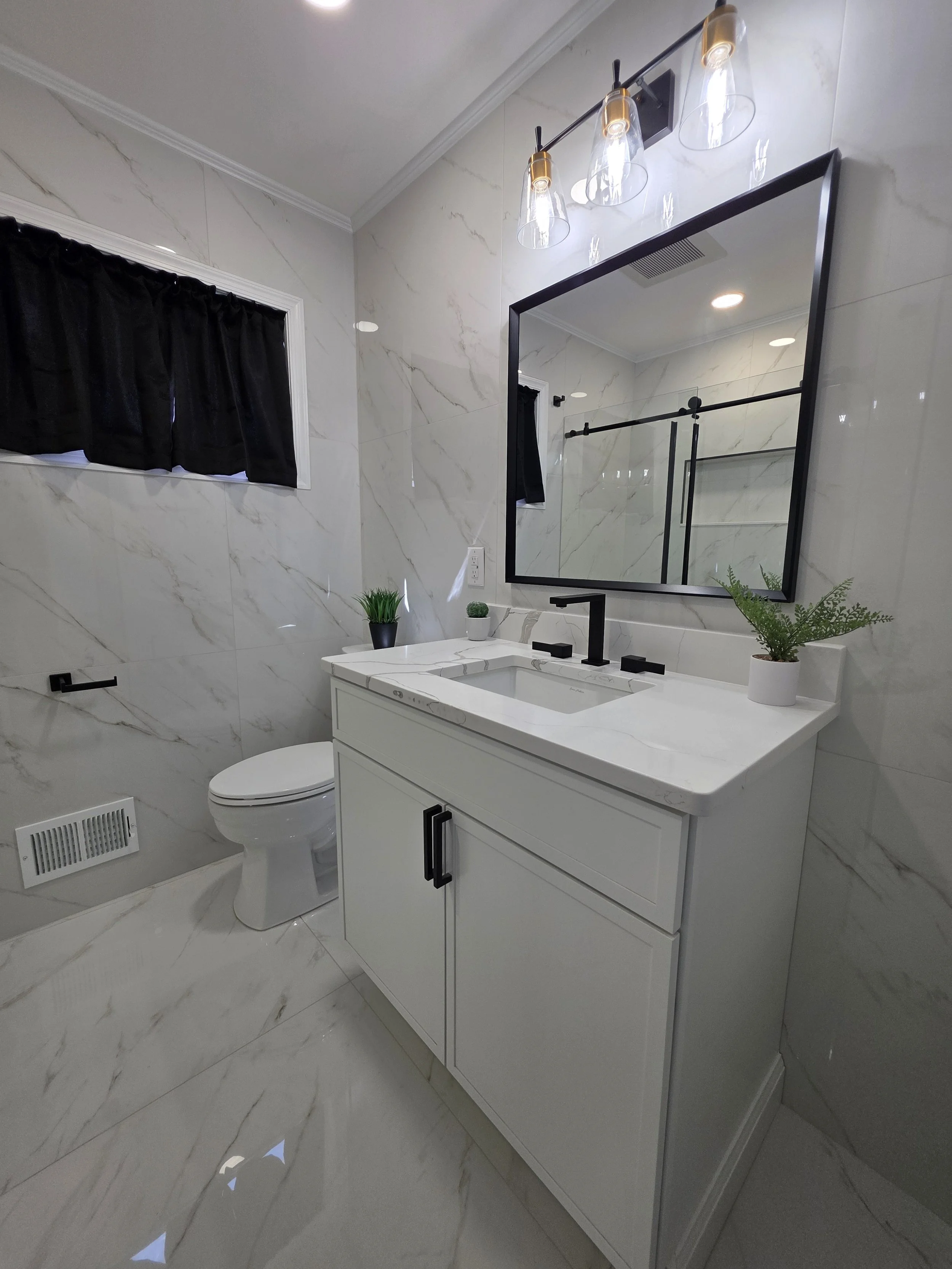 Finished hallway bathroom with a gray vanity, black-framed mirror, black fixtures and marble-look tile around the toilet and window.