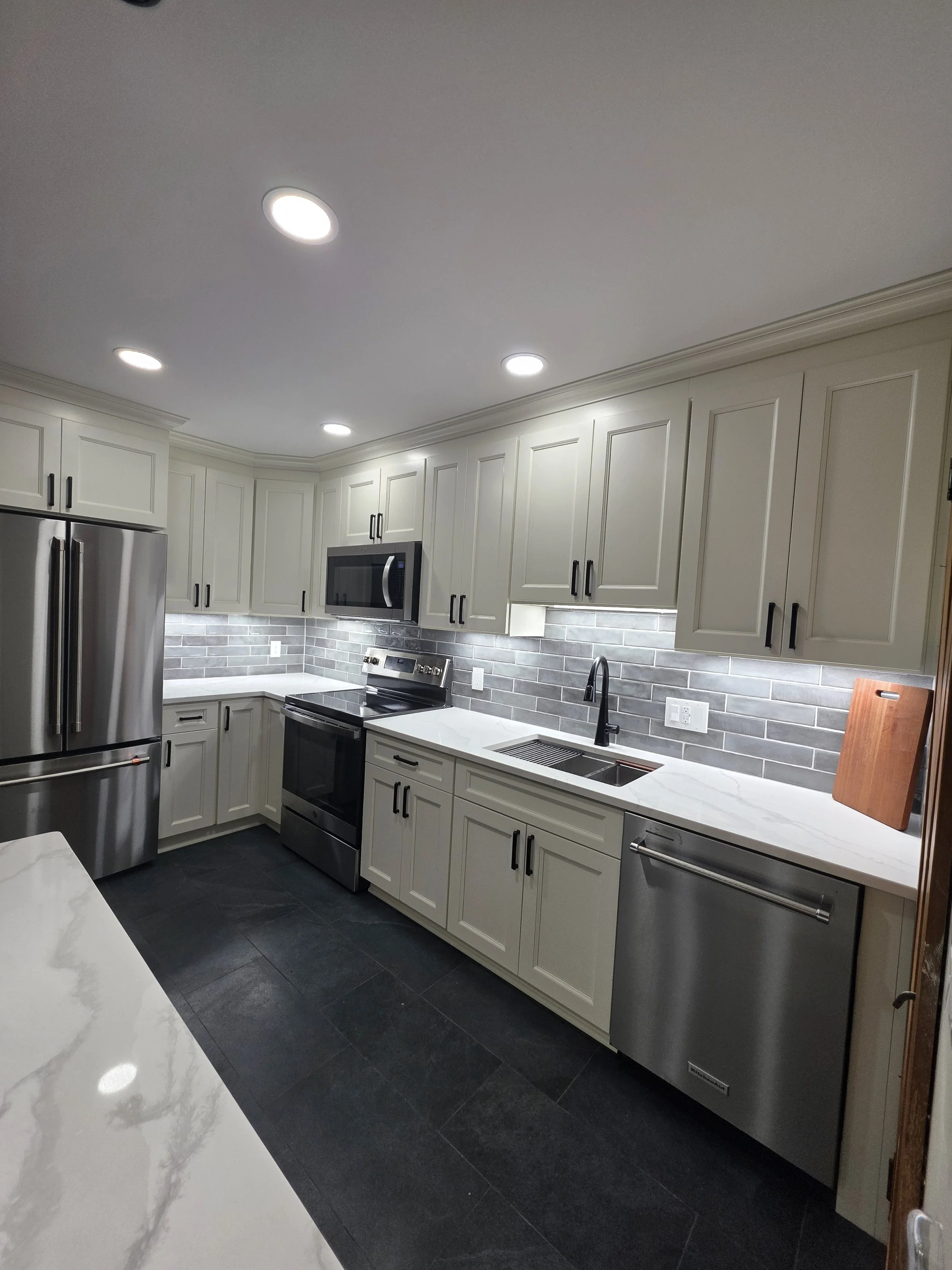 Wide view of the kitchen with light cabinetry, stainless appliances, gray backsplash and dark floor tile.