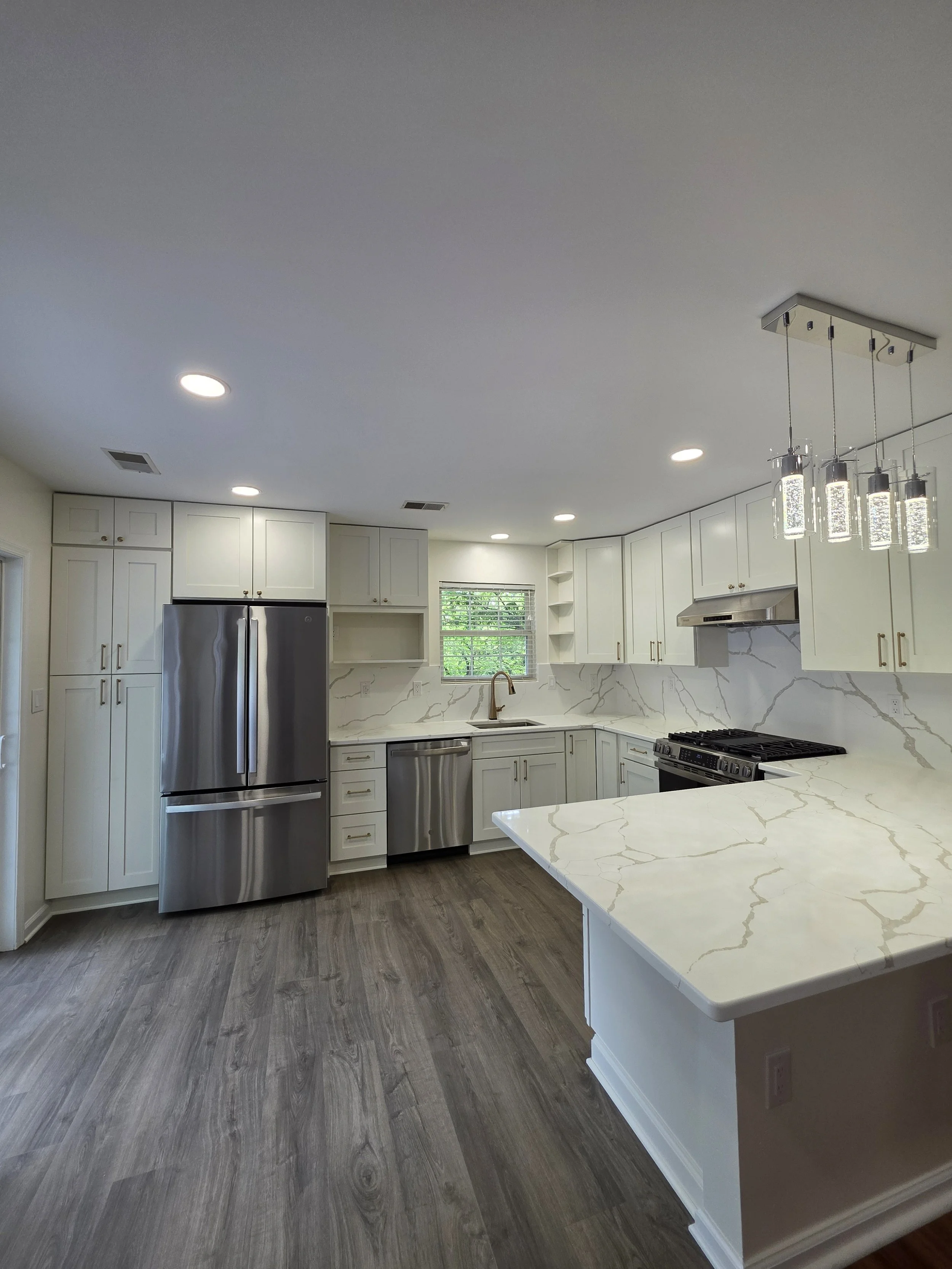 Wide view of Sameer and Shital’s remodeled white kitchen with a veined peninsula and wood-look flooring.