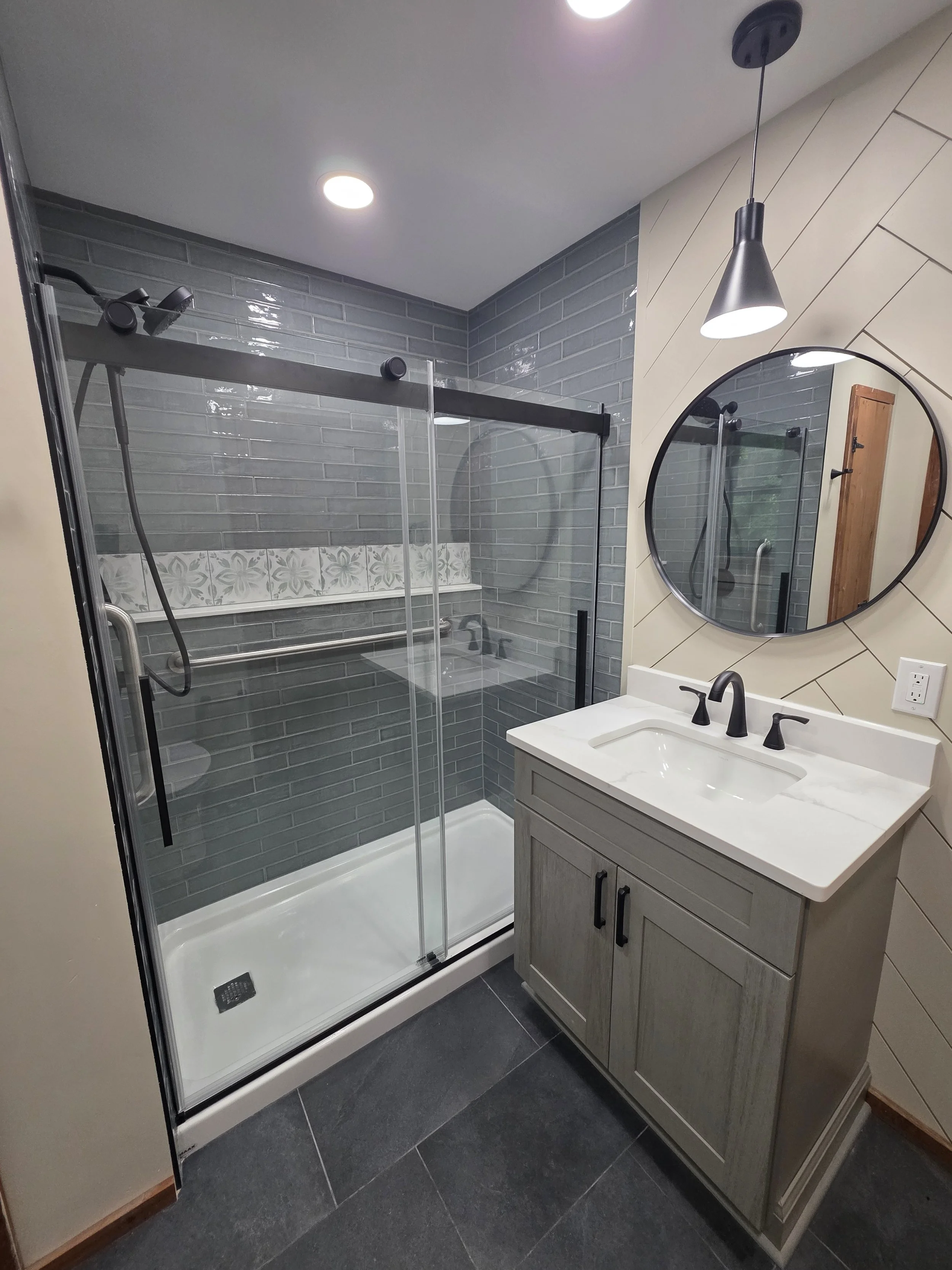 Donna and Chris’s remodeled master bathroom with a glass shower, wood-look vanity, round mirror and dark fixtures.
