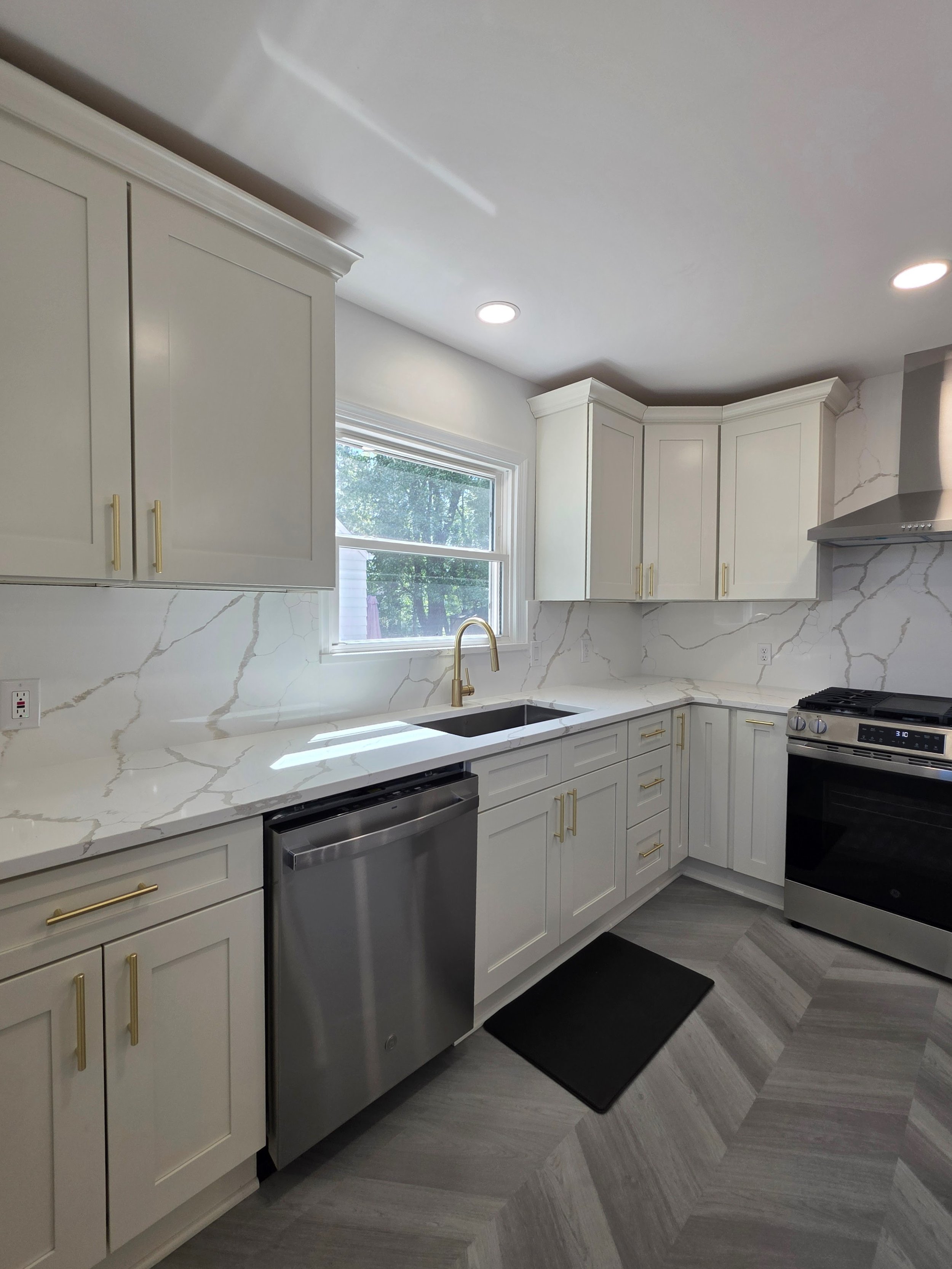 Sink beneath the window with white cabinetry, dishwasher and herringbone-pattern flooring.