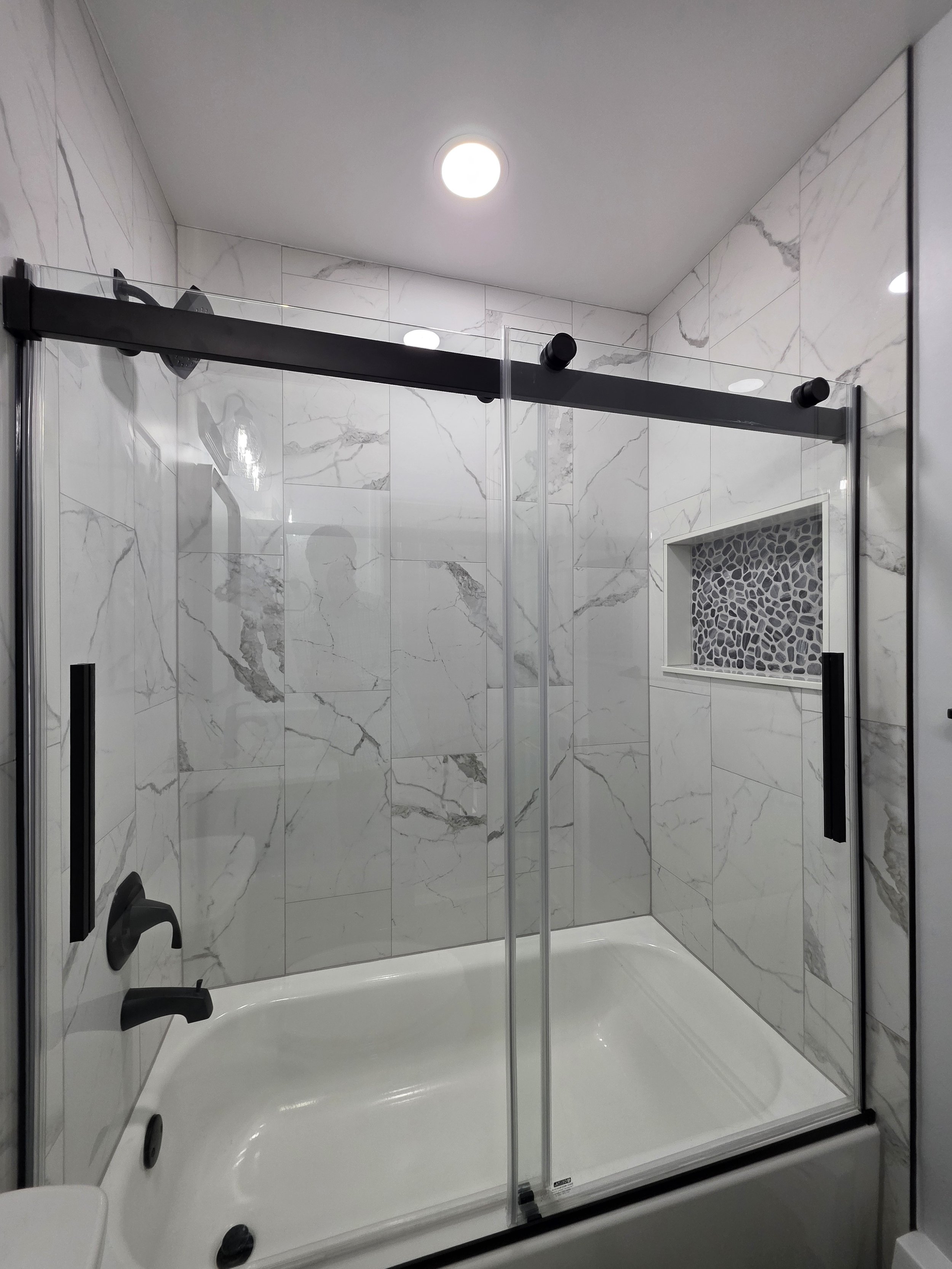Glass tub enclosure with black sliding-door hardware and marble-look wall tile.