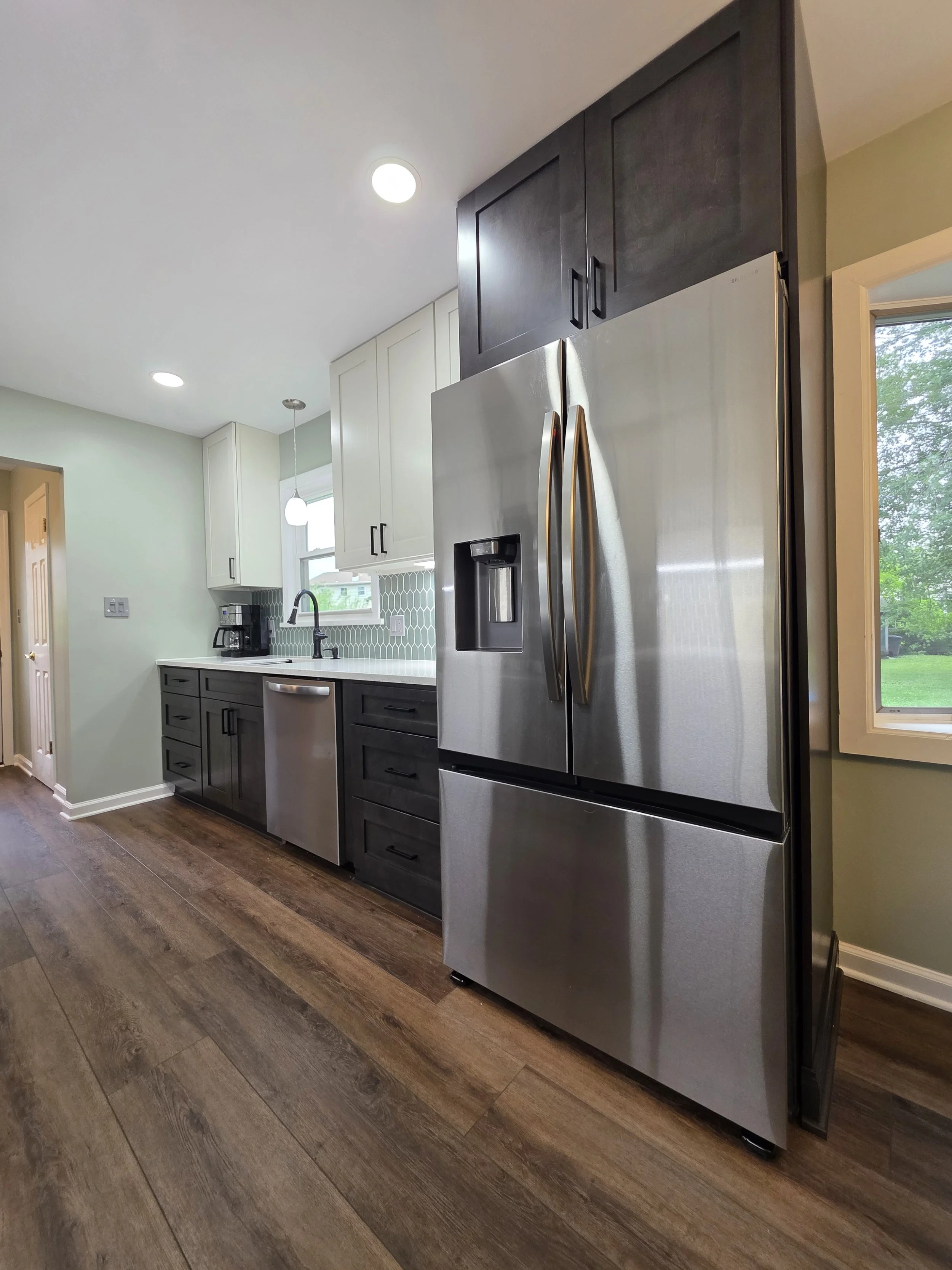 Wide view of the refrigerator and sink wall with contrasting upper and lower cabinetry.