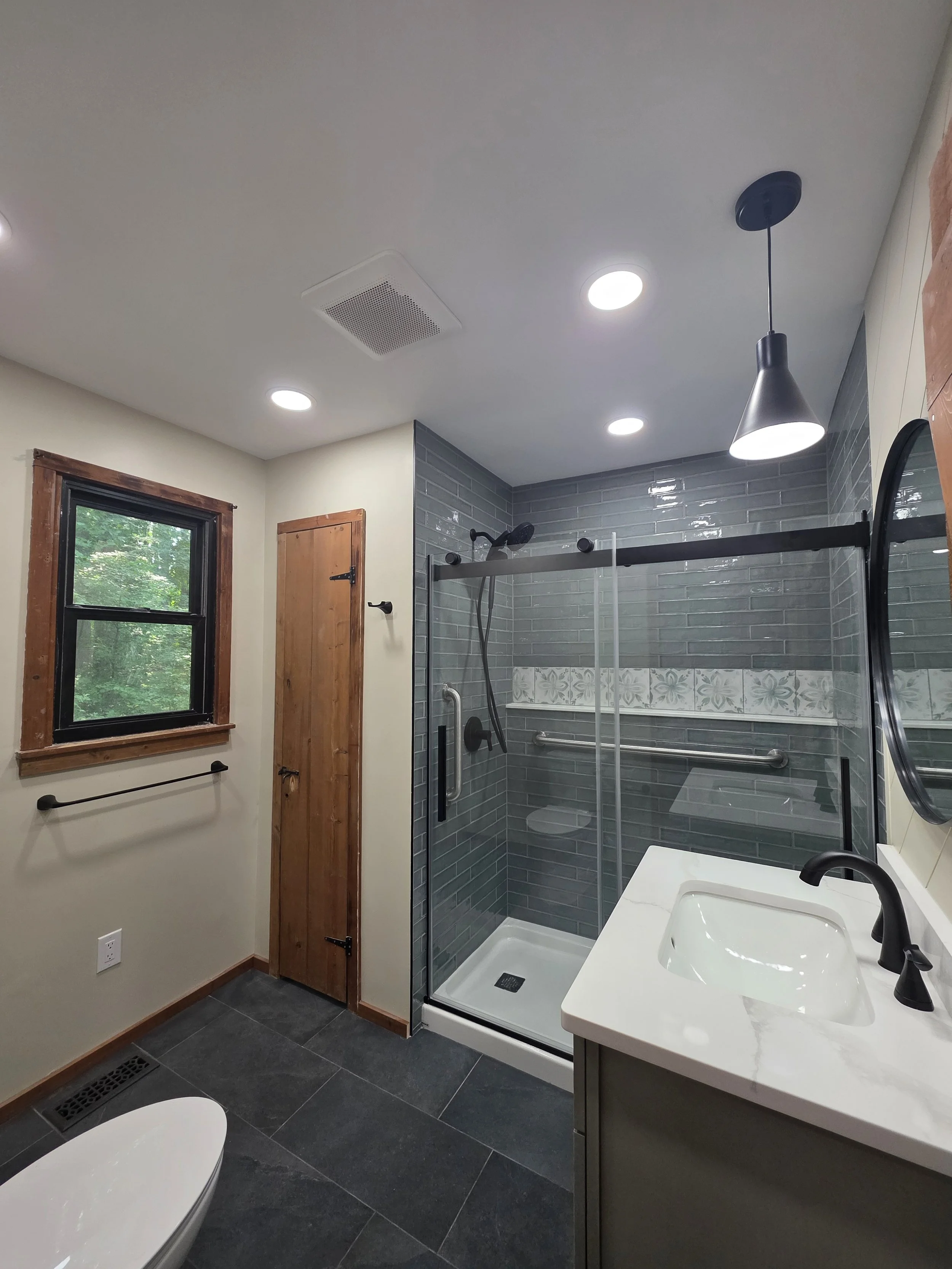 Wide view of the remodeled bathroom with dark floor tile, recessed lights and a glass shower enclosure.