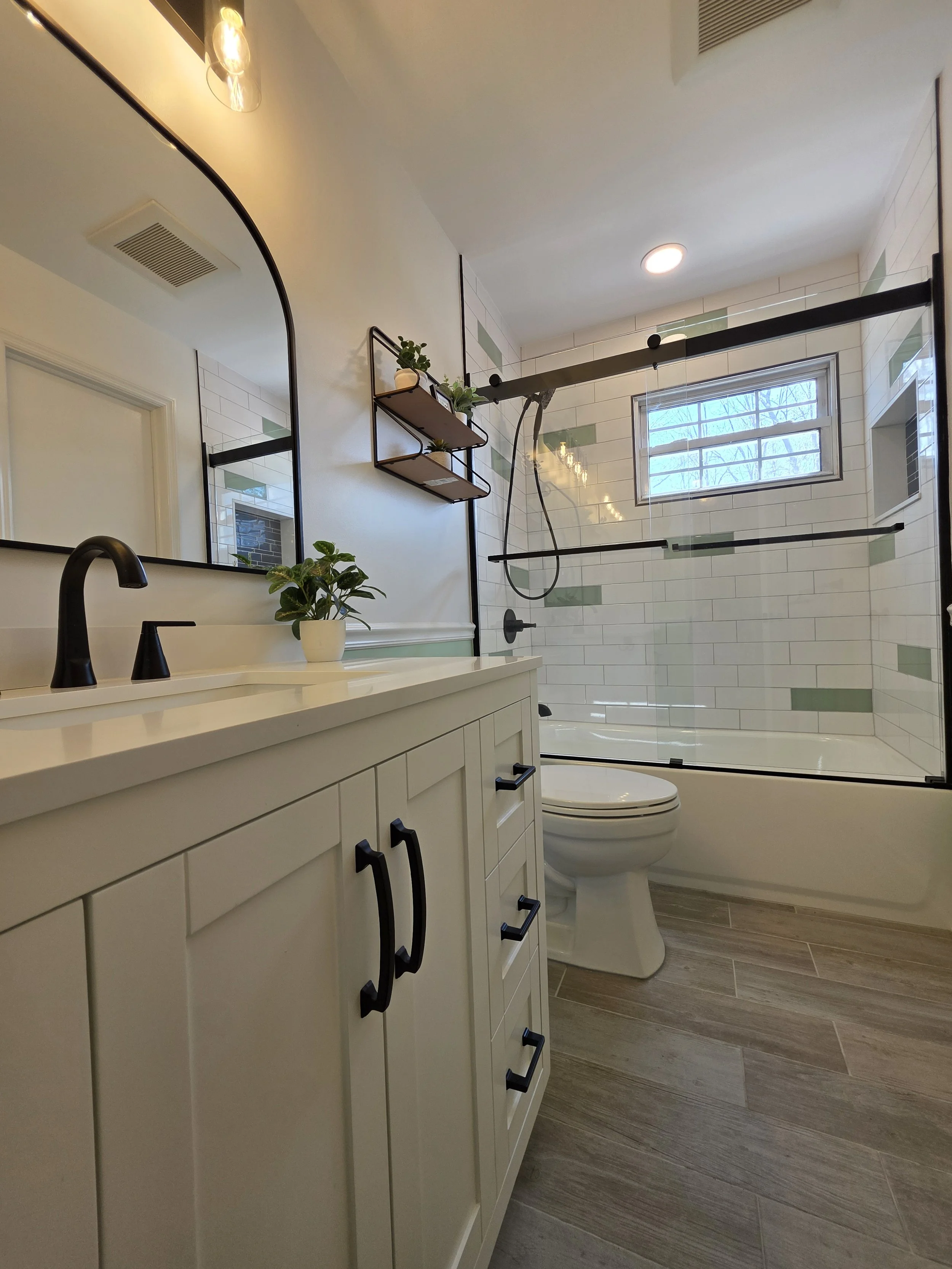 White vanity with black faucet and rounded mirror beside the toilet and remodeled bathtub.