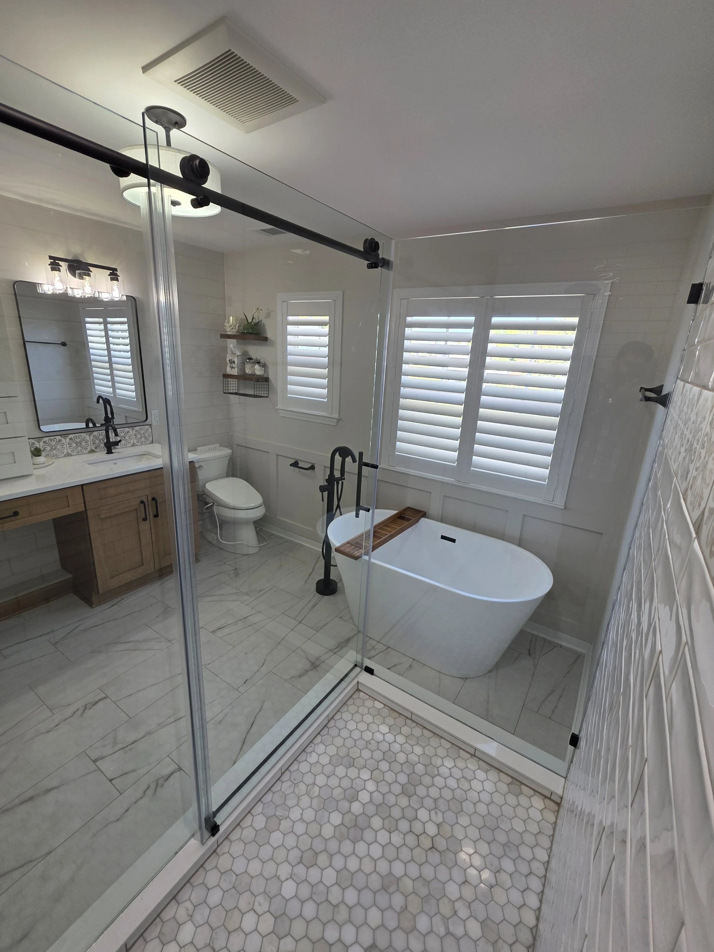 View from the tiled shower toward the freestanding tub, vanity and shuttered windows.