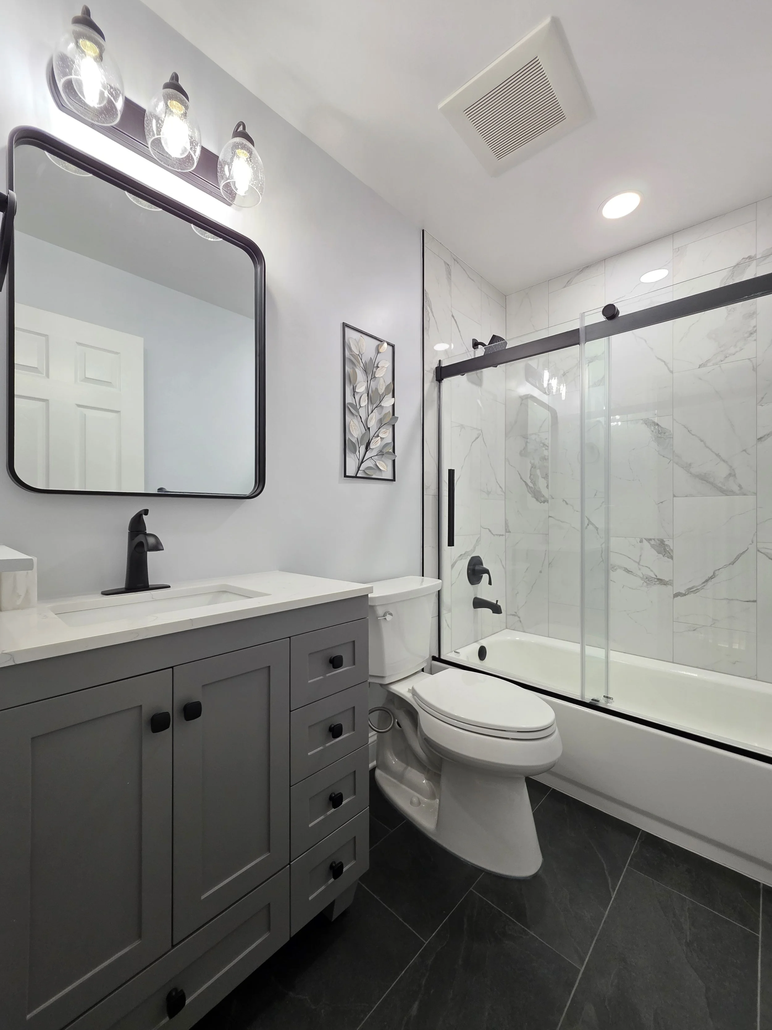 Liz’s remodeled hallway bathroom with a gray vanity, black fixtures, dark floor tile and a glass tub enclosure.