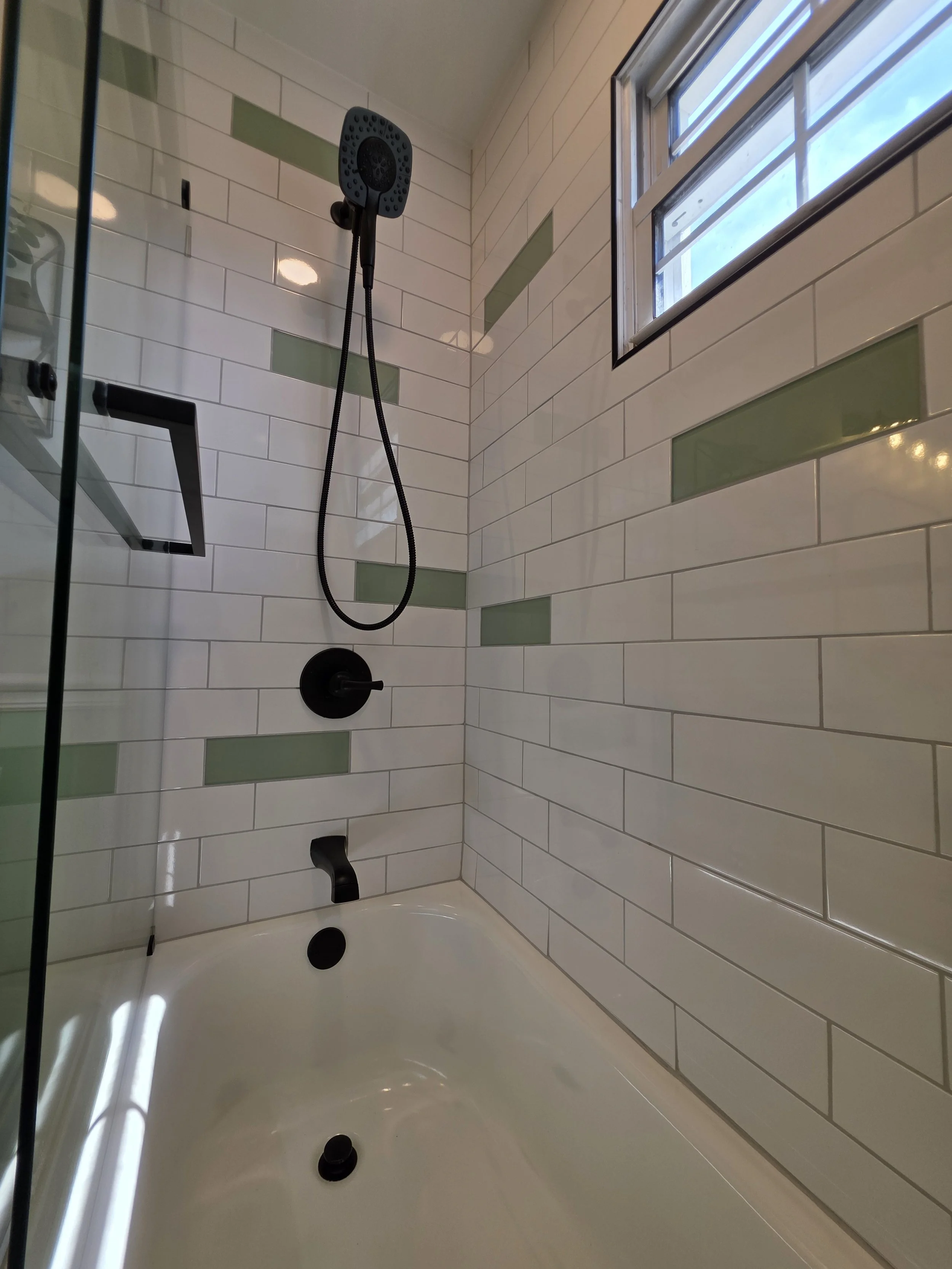 Black shower fixtures and handheld spray above the white bathtub, surrounded by white tile with green accents.