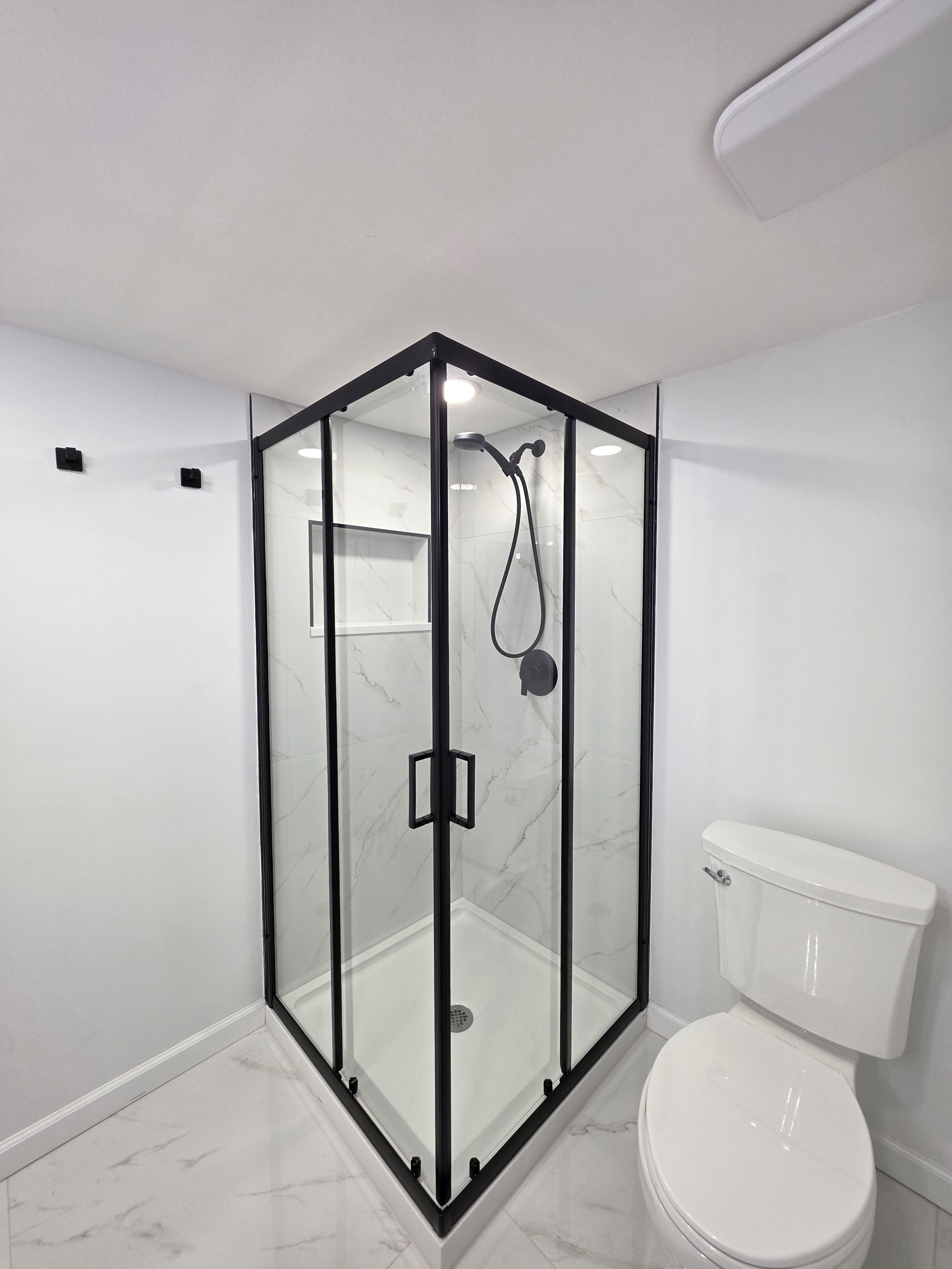 Corner shower enclosure beside the toilet, with black framing and marble-look walls and floor.