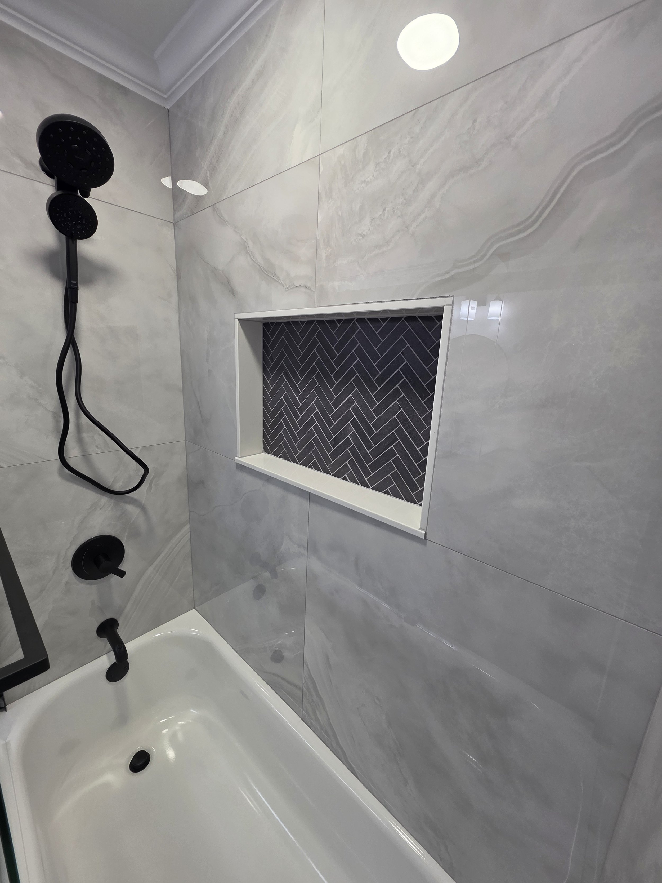 White bathtub beneath black shower fixtures and marble-look wall tile with a dark herringbone niche.