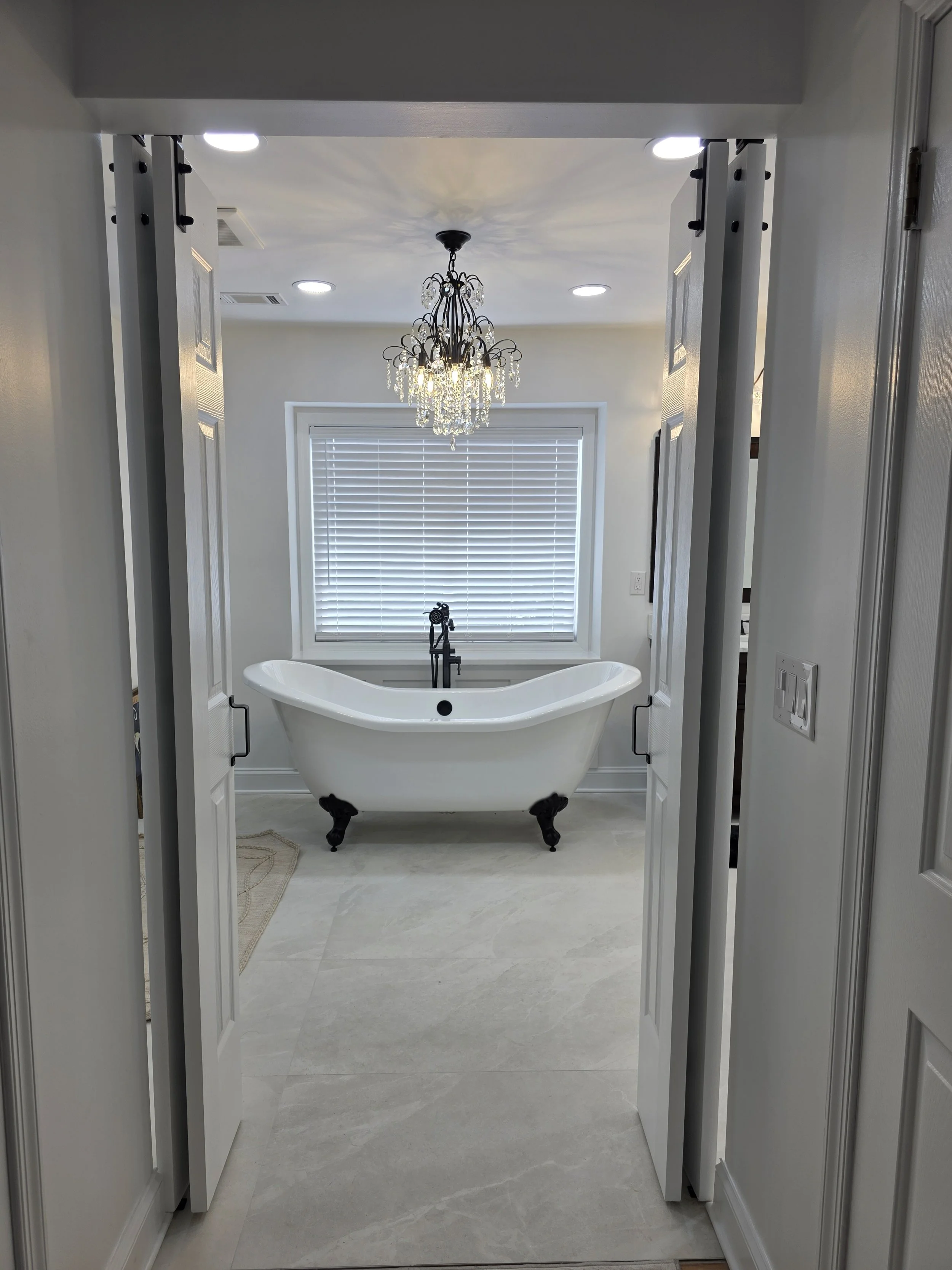 View through the bathroom doorway toward the freestanding clawfoot tub and chandelier beneath a large window.
