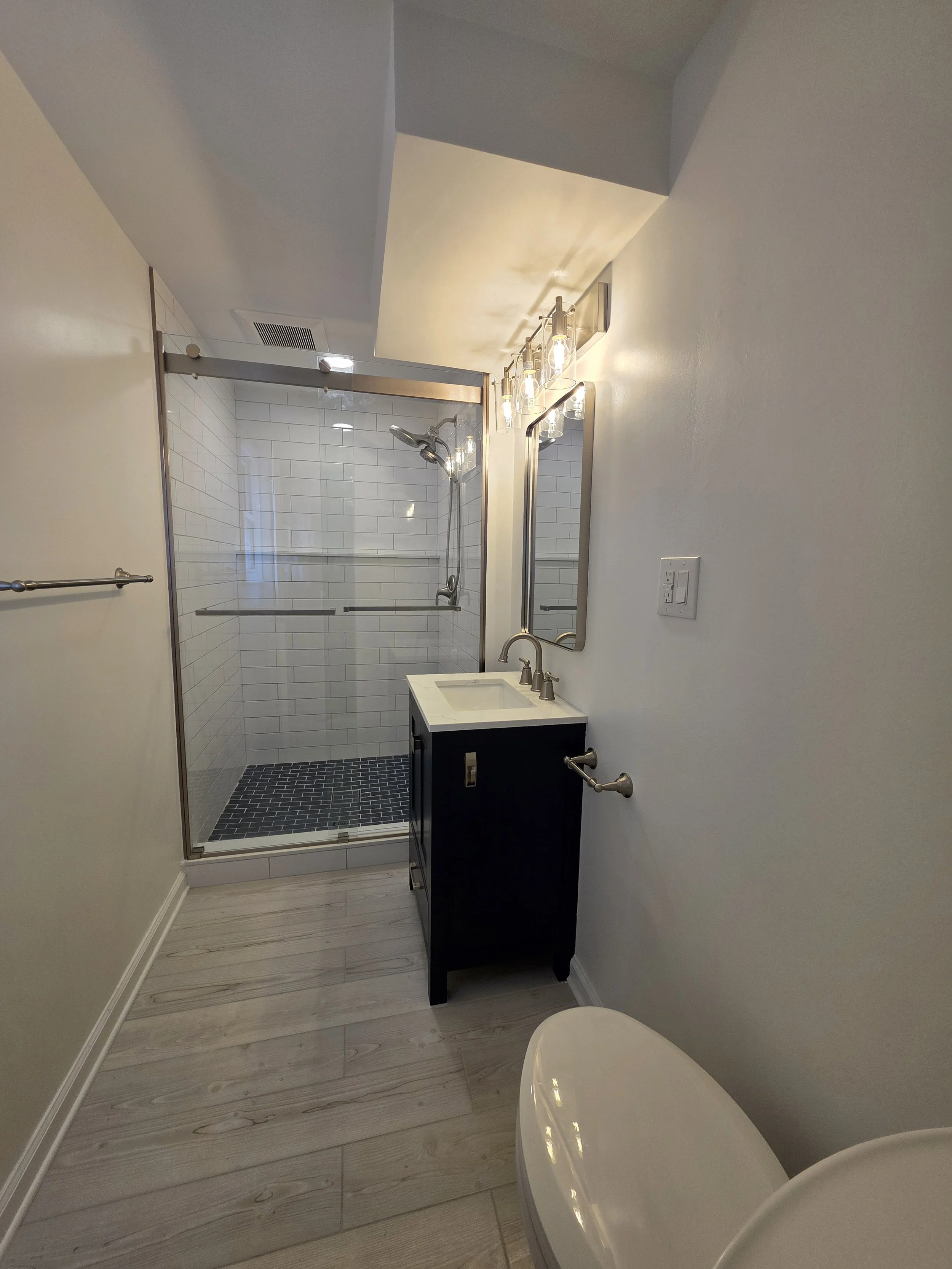 Wide view through the basement bathroom toward the glass shower, with navy vanity and light wood-look floor.