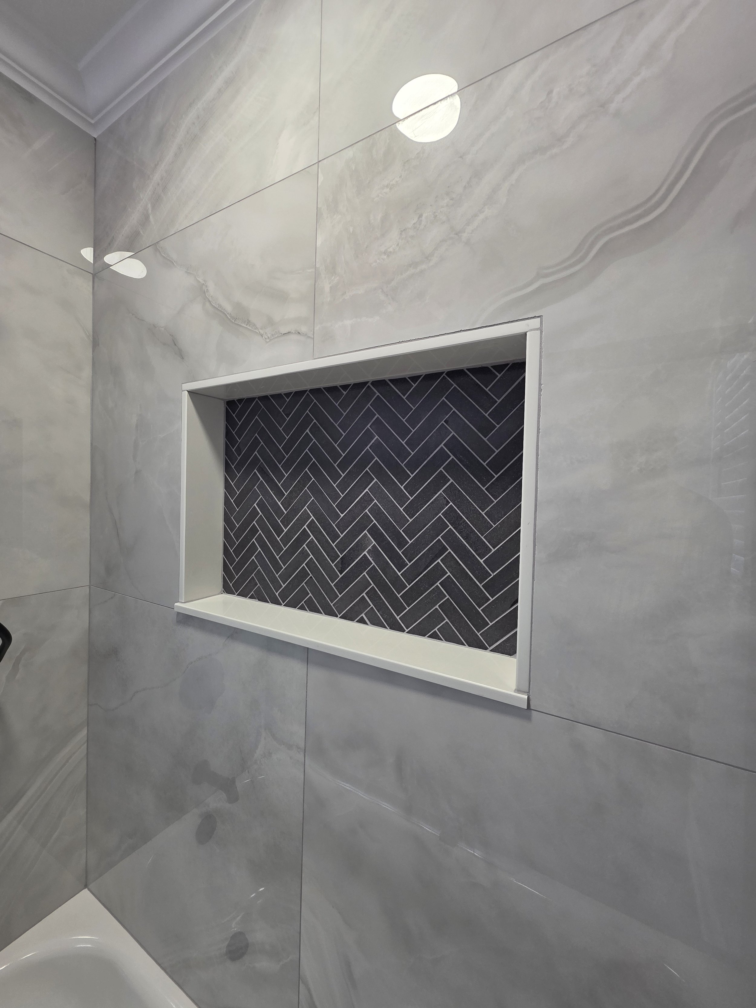 Close view of the recessed niche with dark herringbone tile inside the marble-look tub surround.