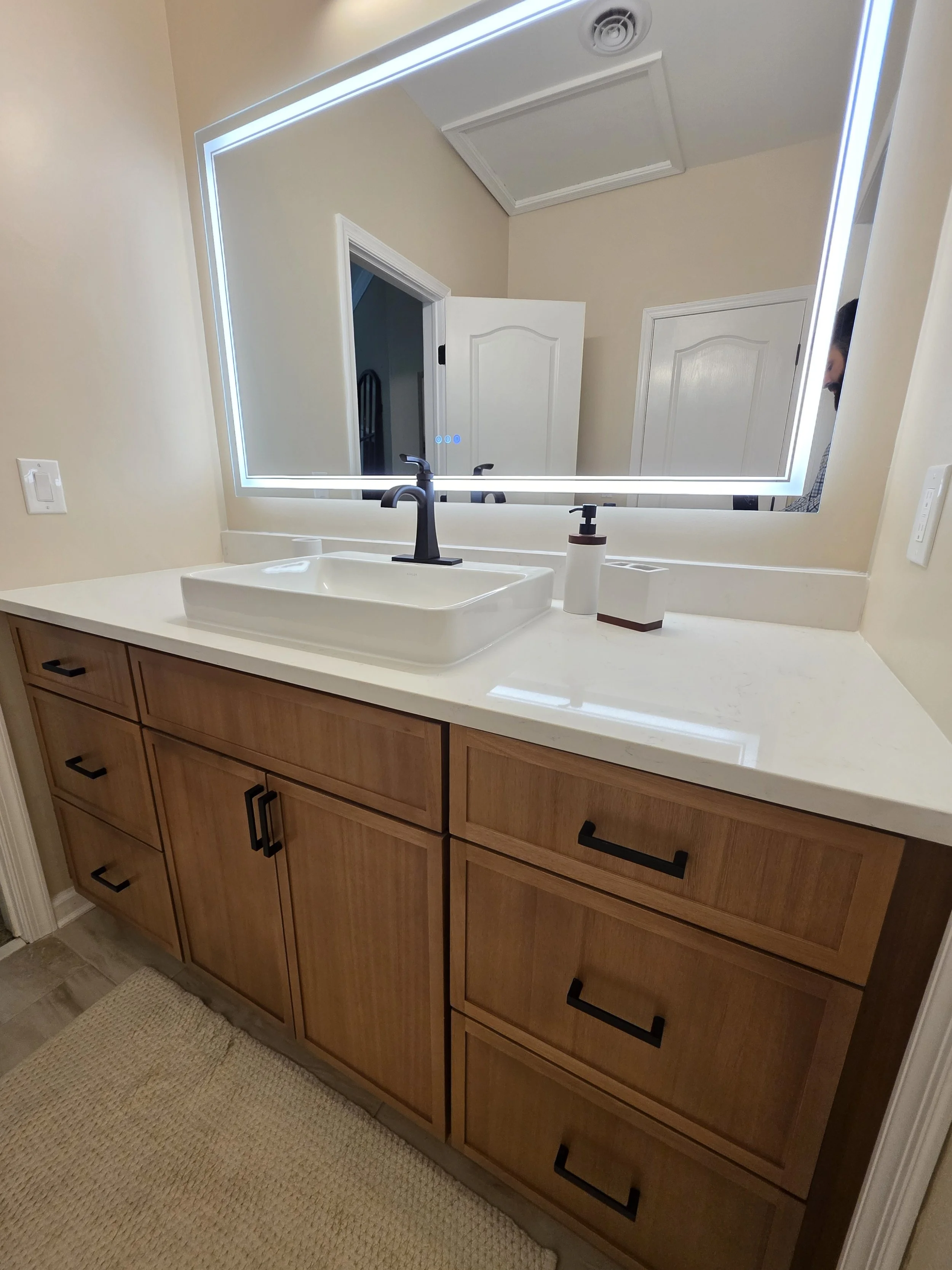 Closer view of the vanity sink, black faucet and illuminated mirror above warm wood-tone cabinetry.