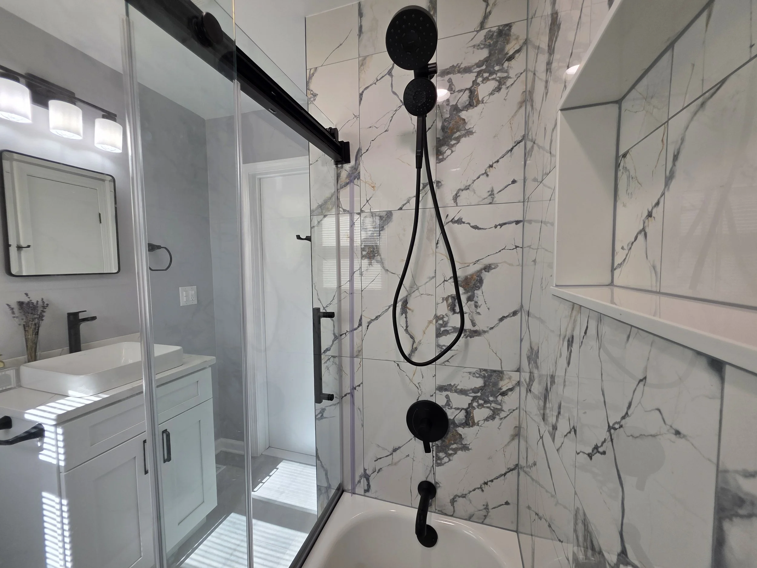 View from the bathtub toward the white vanity, with black shower fixtures and veined tile walls.