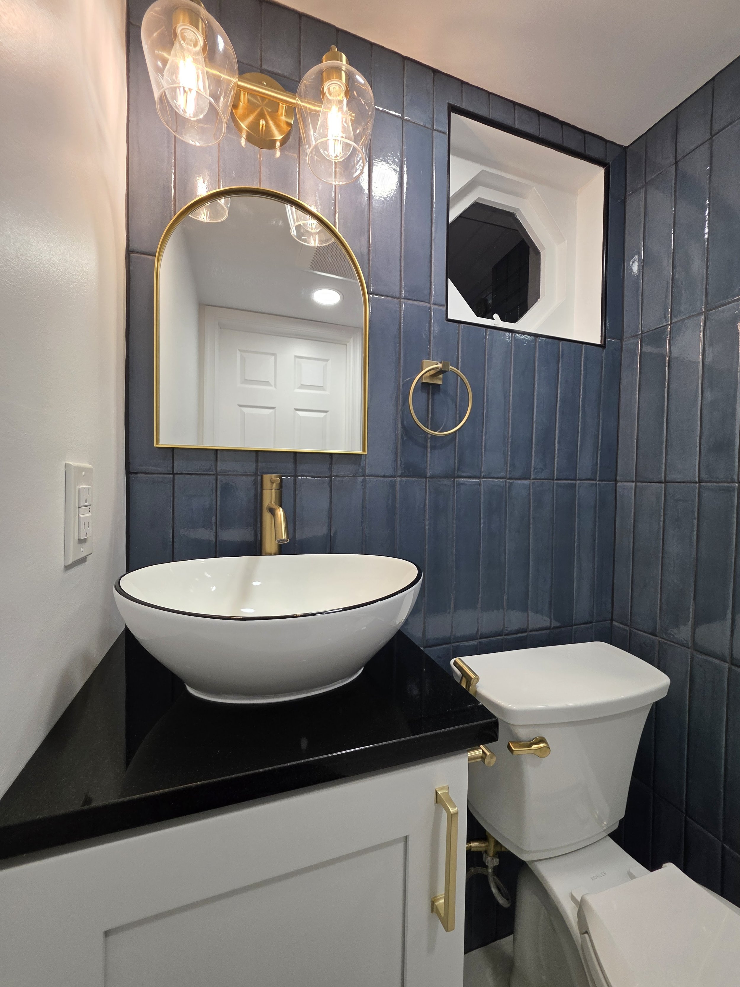 White vessel sink on a dark countertop, with a gold-tone faucet, arched mirror and blue wall tile.