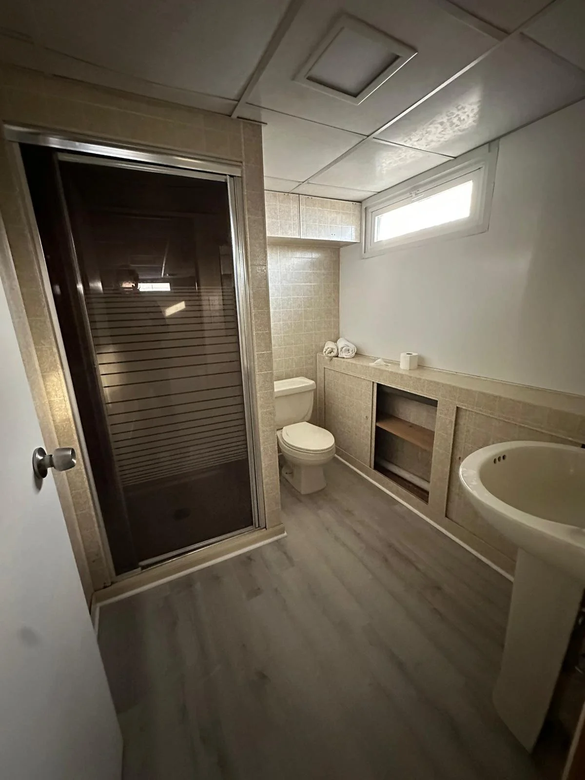 Before renovation: the original basement bathroom with a framed shower, pedestal sink and older wall and ceiling finishes.
