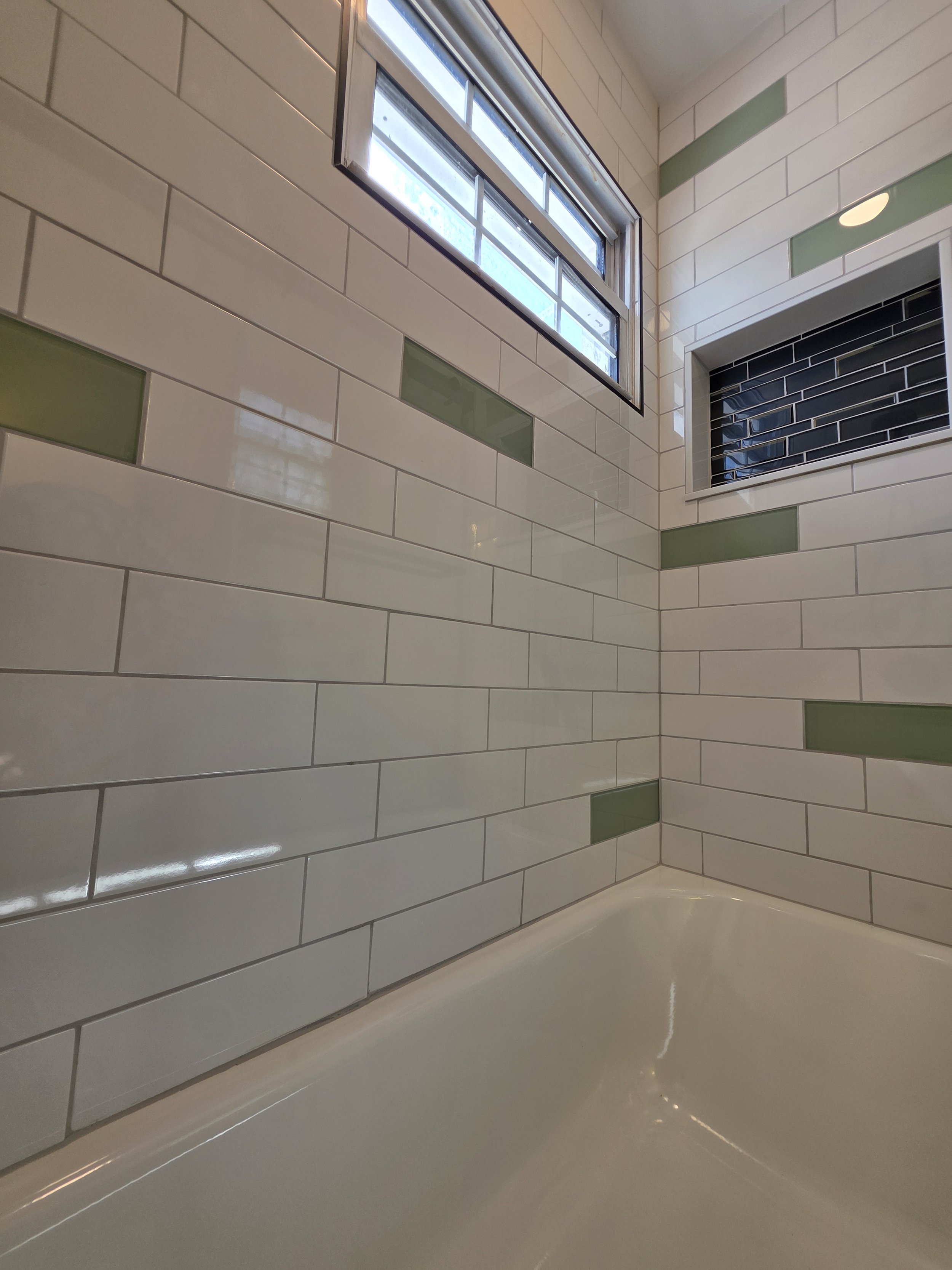 White tub surround with scattered green accent tiles, a dark recessed niche and a high window.