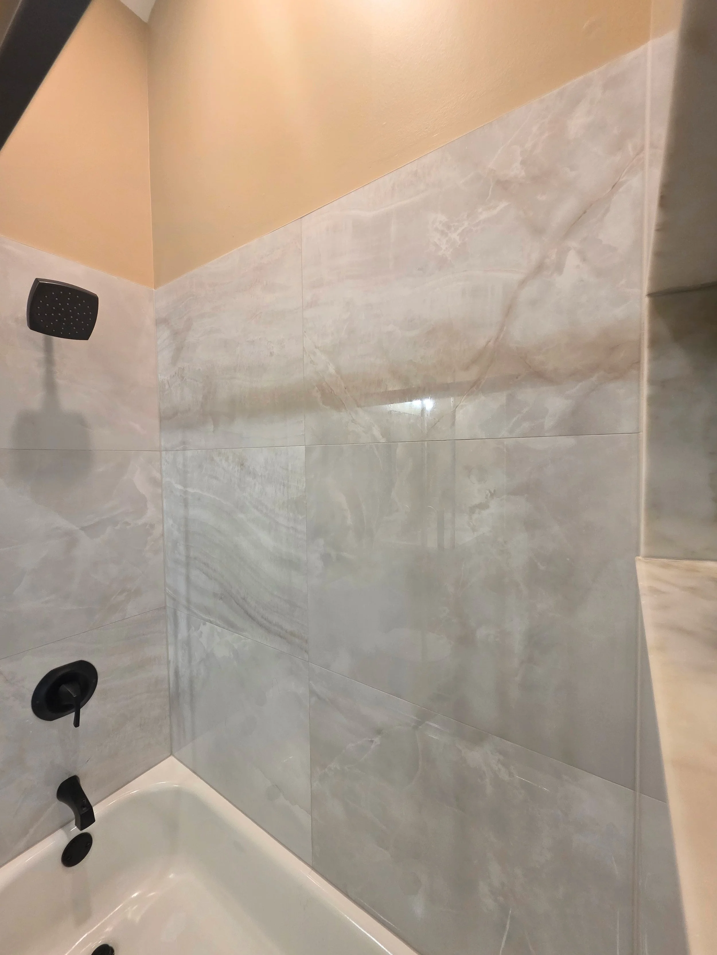 Black shower fixtures above a white bathtub surrounded by light veined wall tile.