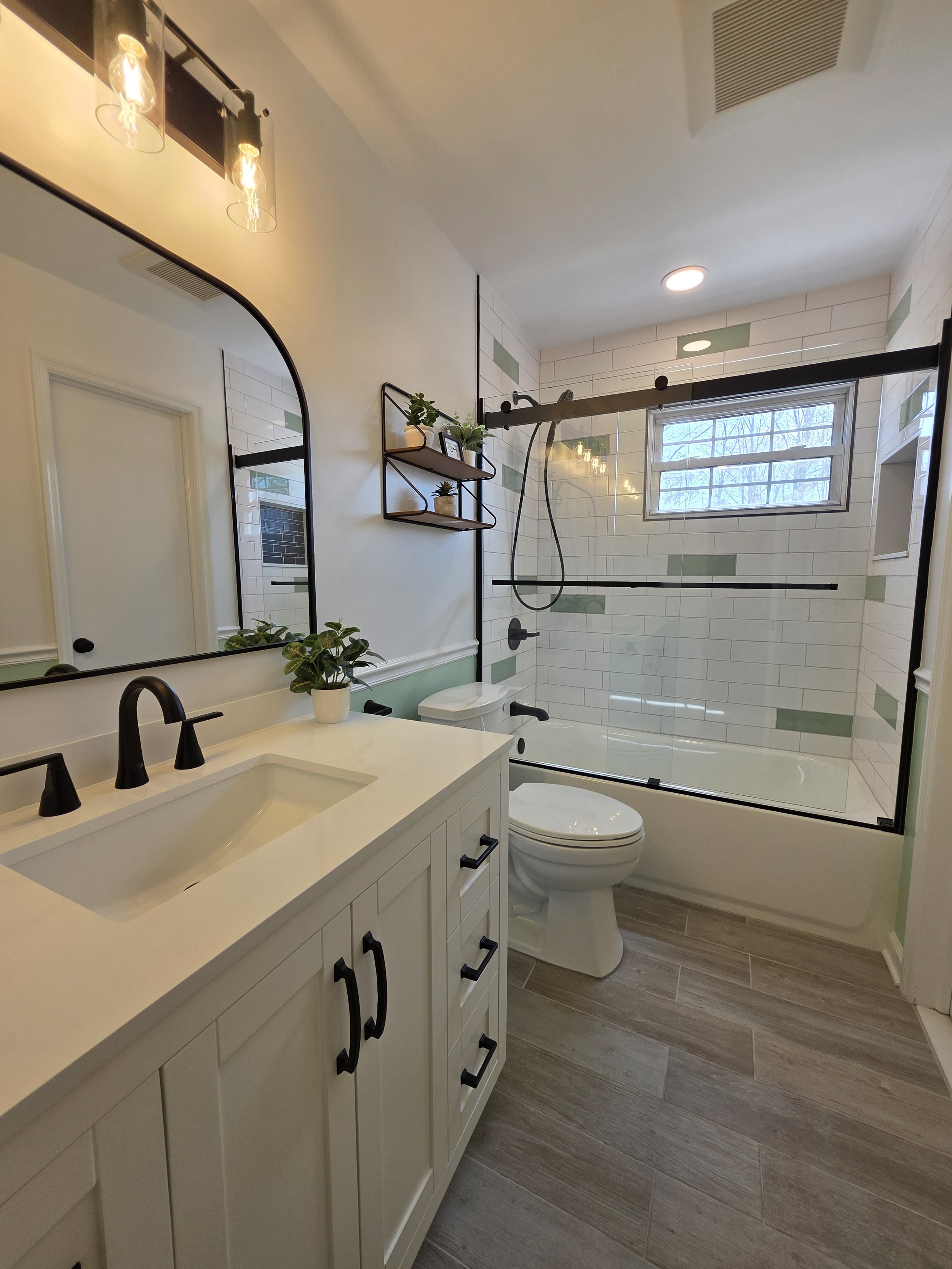 Finished hallway bathroom with white vanity, black fixtures, green tile accents and a glass tub enclosure.