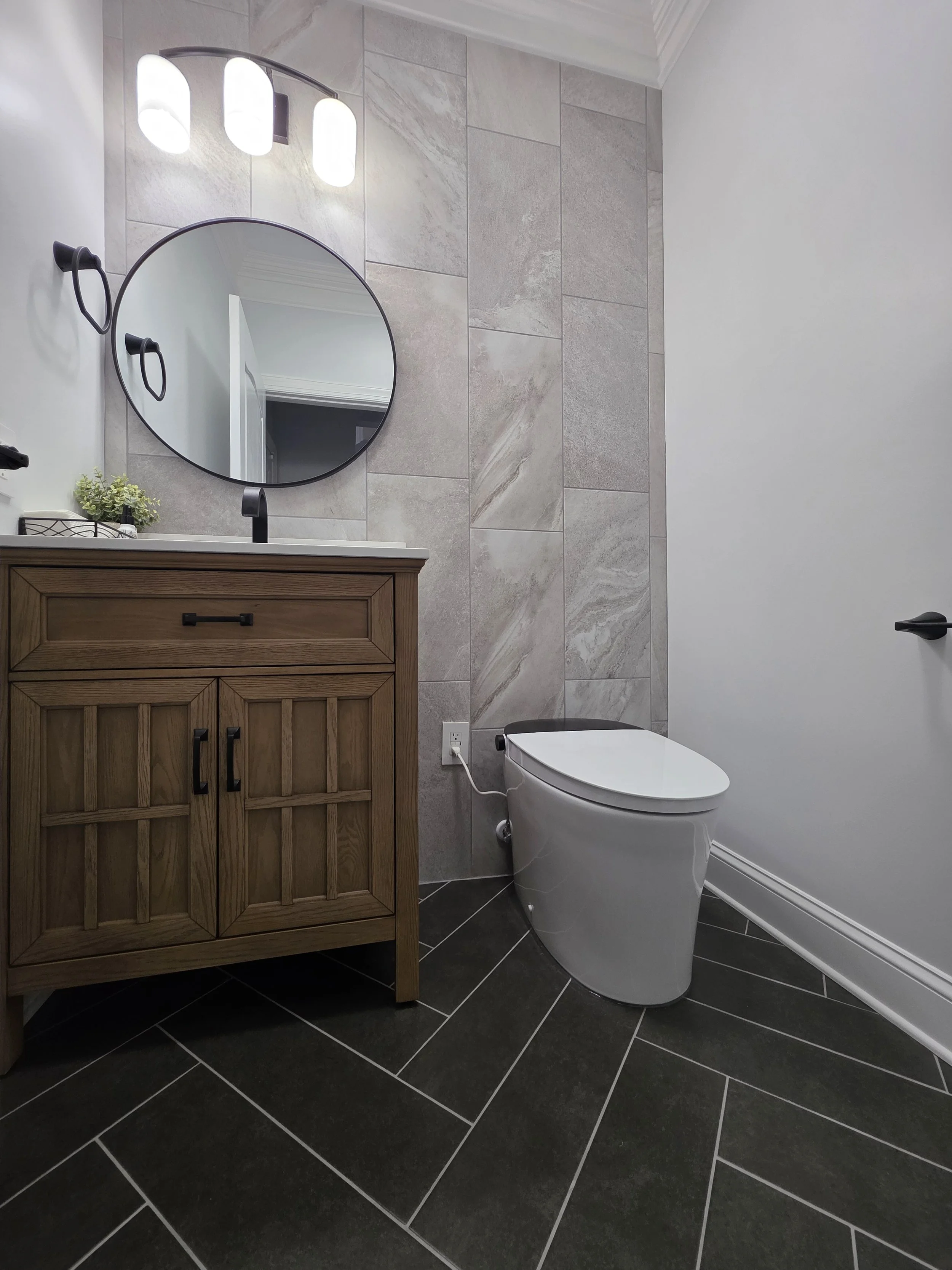 Low view of the vanity and toilet against the tiled accent wall above dark herringbone floor tile.