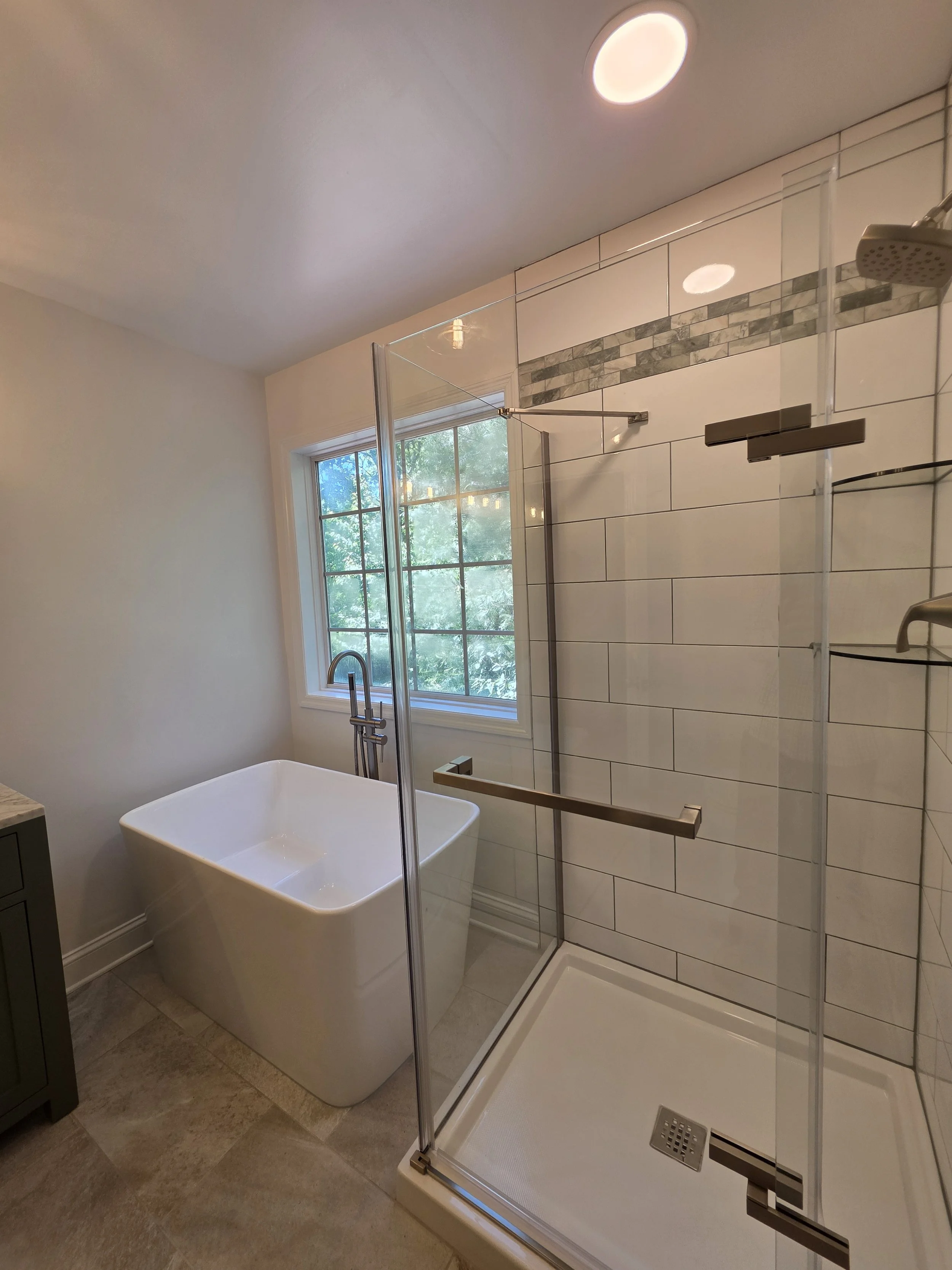 Glass shower enclosure with white wall tile and a decorative accent band beside the freestanding tub.