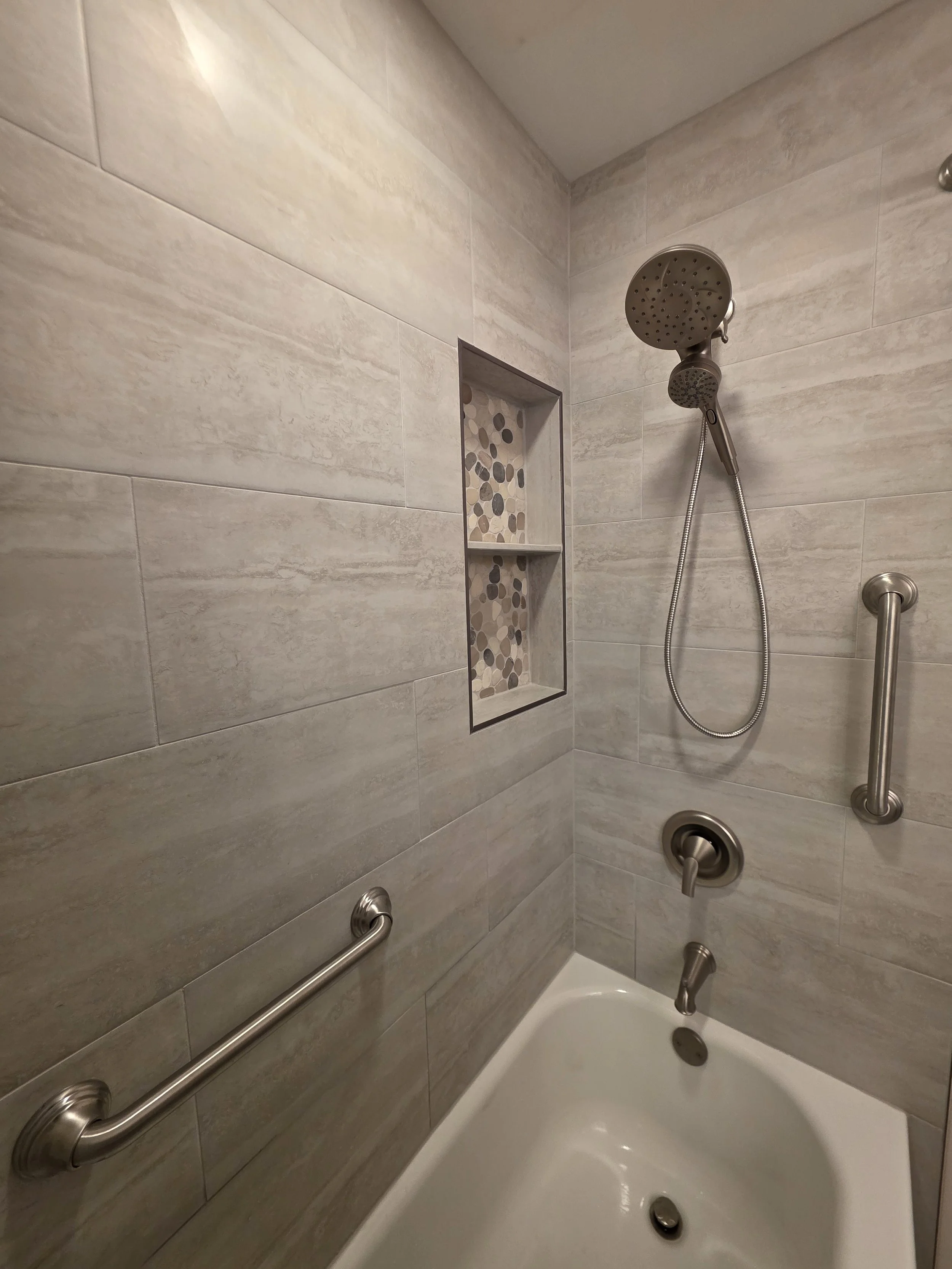 Close view of the tiled tub surround, handheld shower, grab bars and two-part recessed niche.