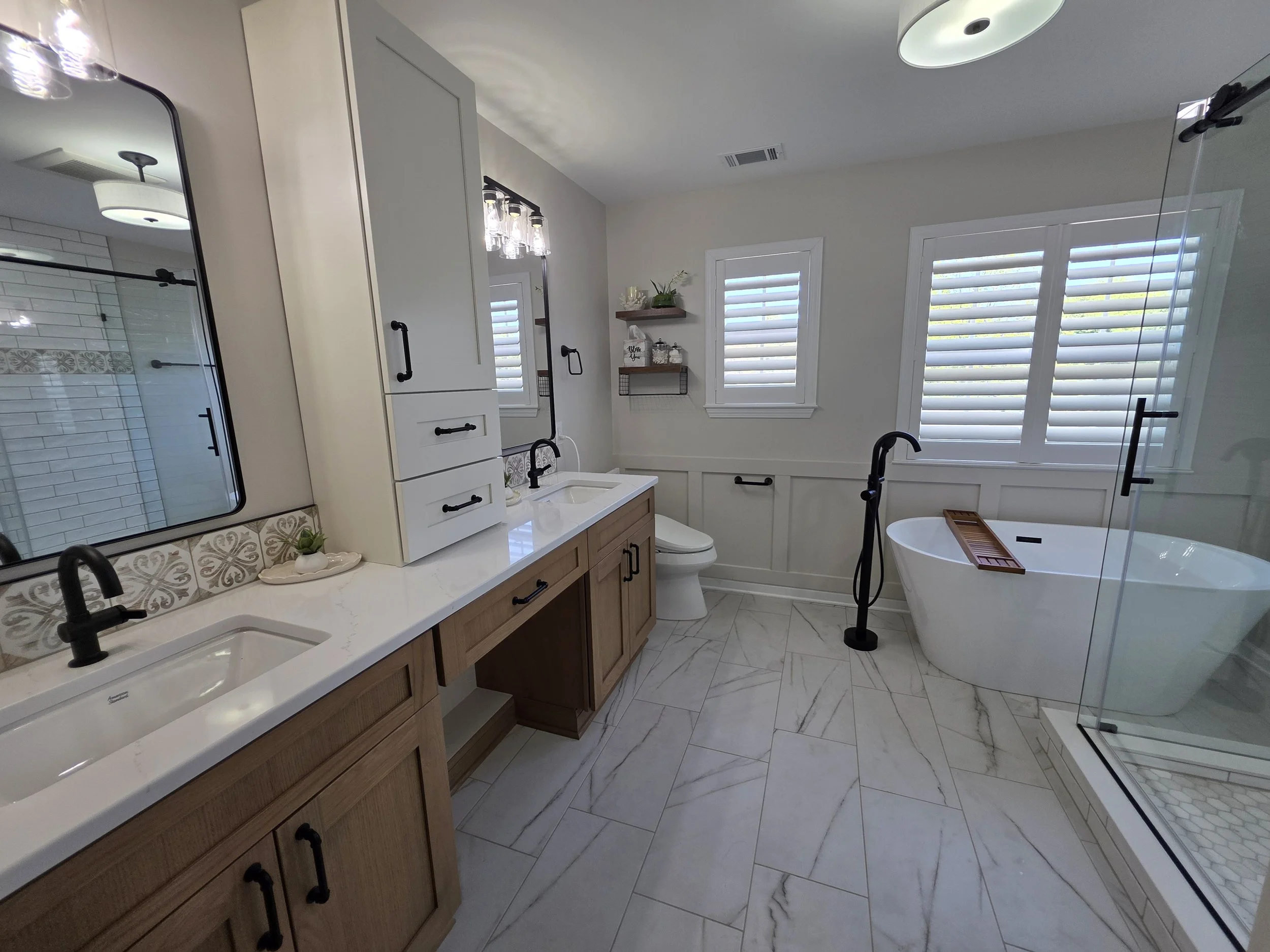 Wide view of Dan and Mohua’s remodeled bathroom with double vanity, tall storage cabinet, freestanding tub and marble-look floor tile.