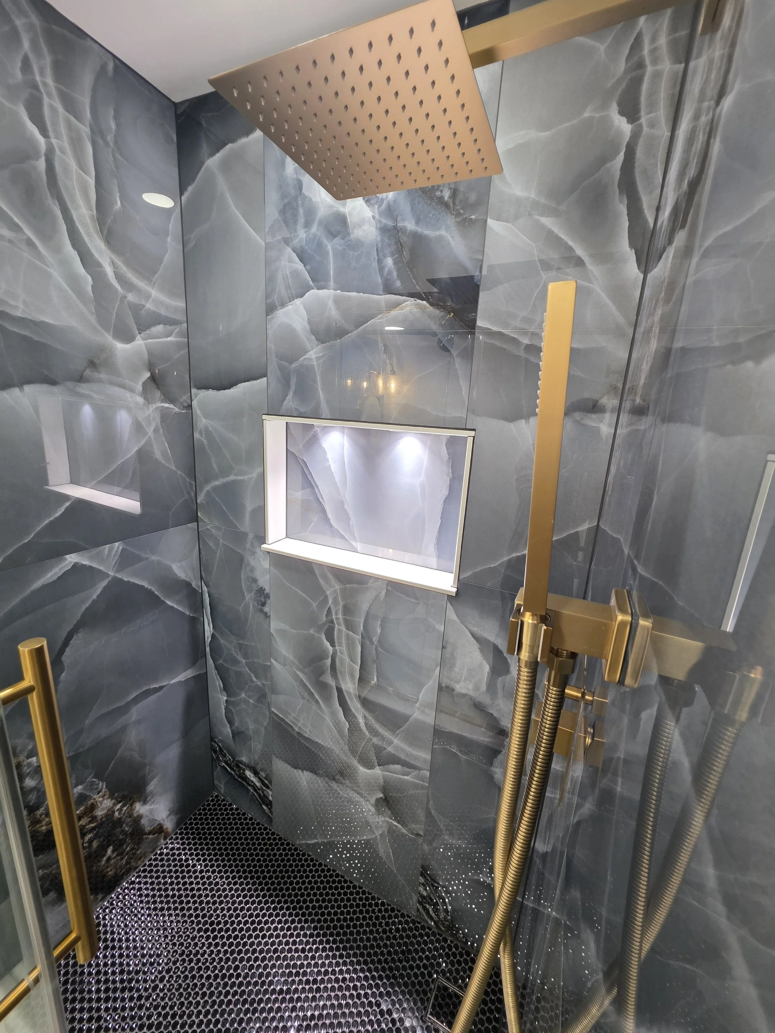 Shower interior with a square rain showerhead, handheld spray, illuminated niche and dark mosaic floor.