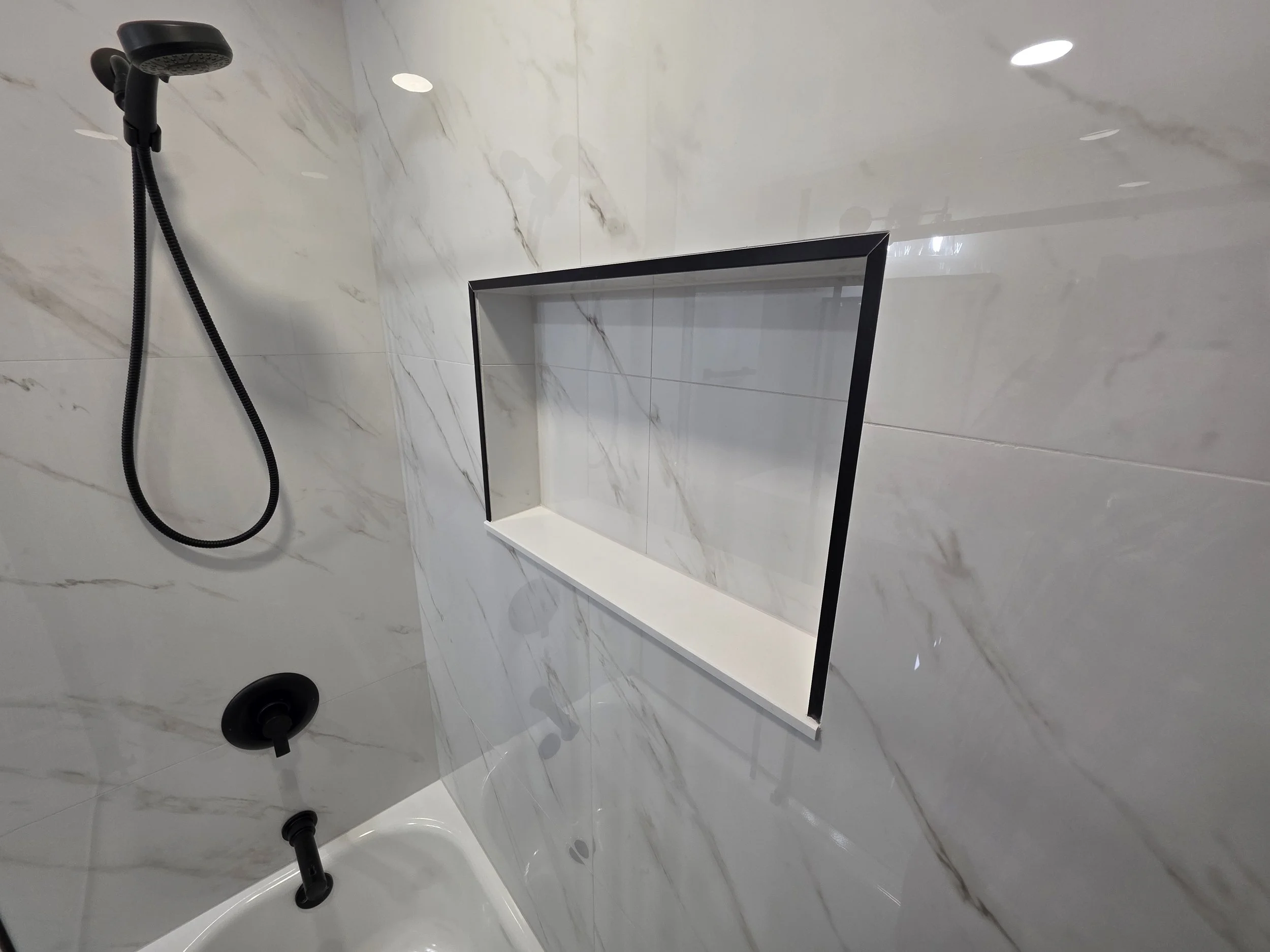 Black tub and shower fixtures beside a wide recessed niche in the marble-look tile surround.
