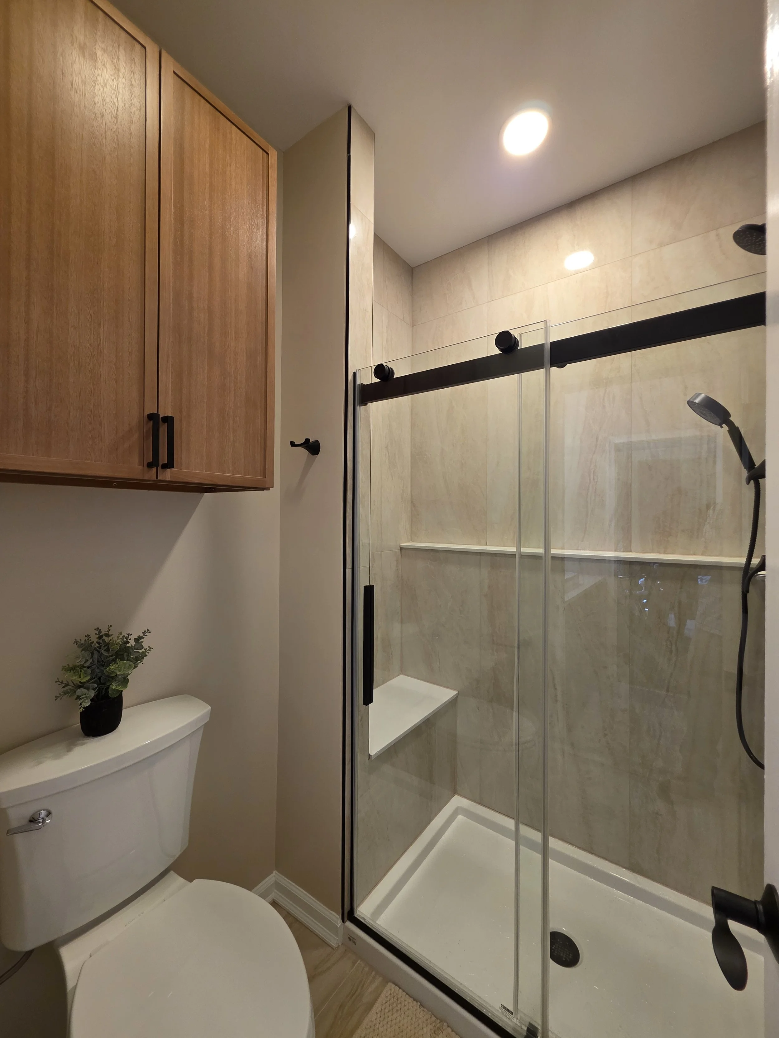 View of the shower enclosure, tiled walls and wood-tone wall cabinet in Rich and Jen’s hallway bathroom.