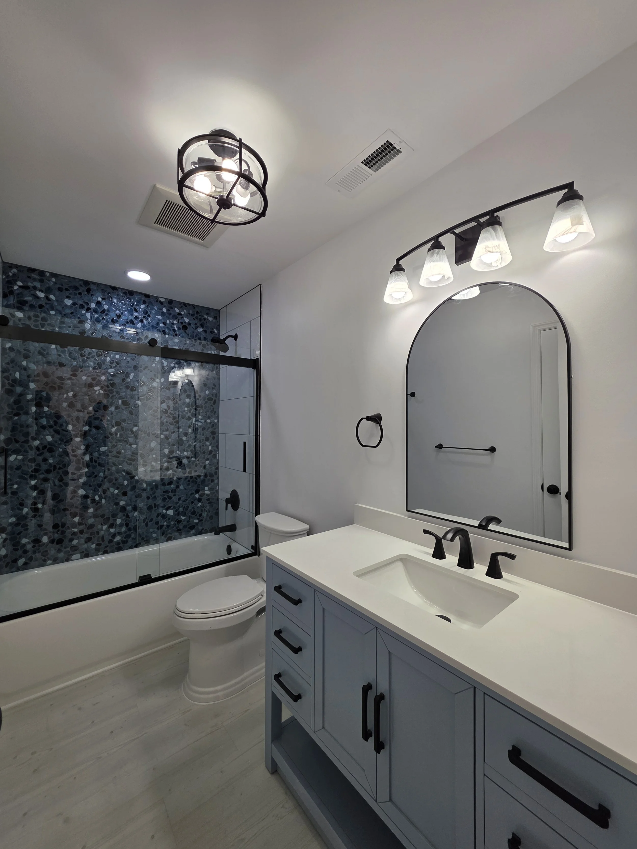 Finished hallway bathroom with a gray vanity, arched mirror, black fixtures and a blue river rock tile accent wall behind the bathtub.
