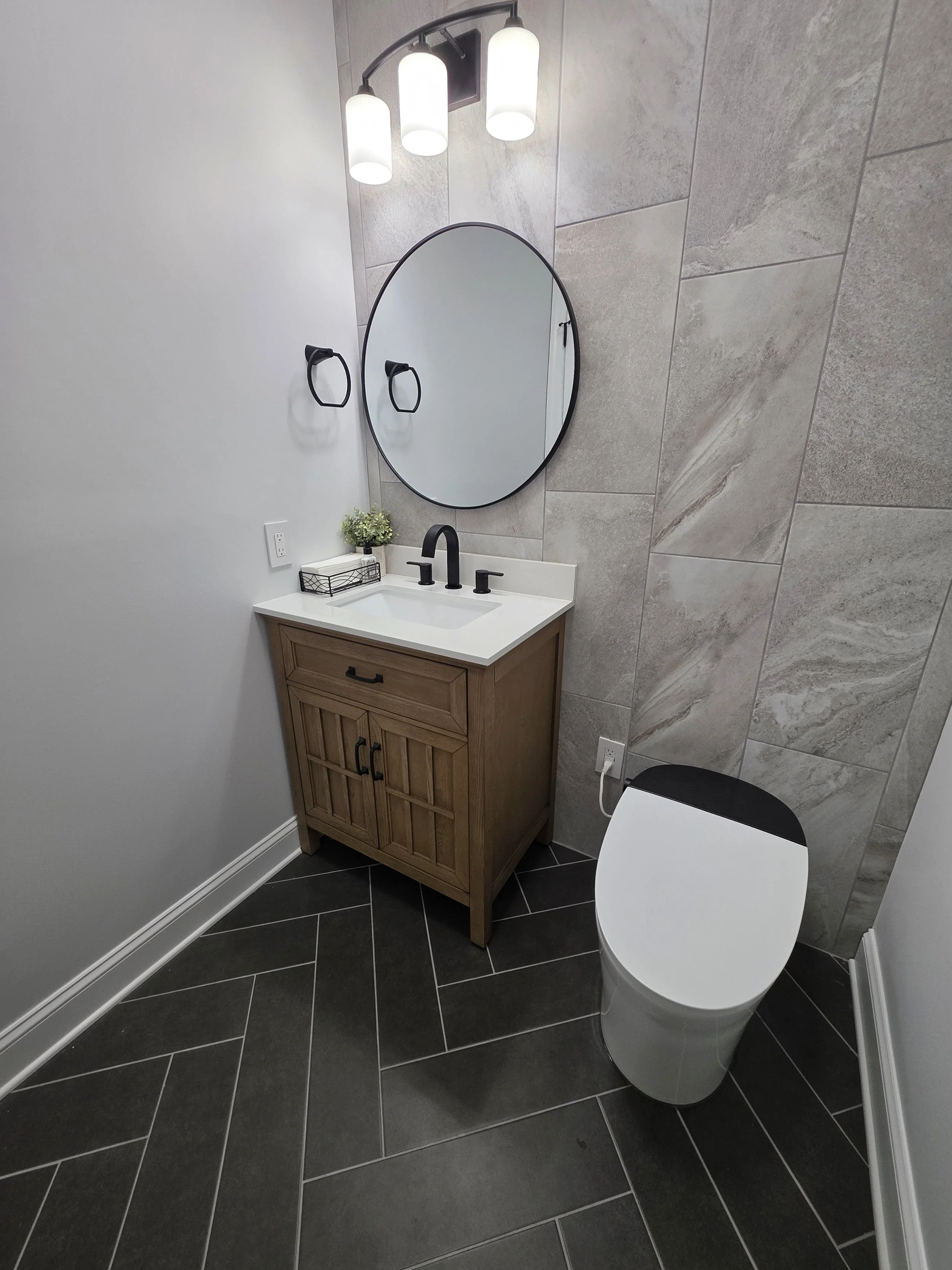 Alan and Alissa’s remodeled powder bathroom with a wood-tone vanity, round mirror, tiled accent wall and dark herringbone floor.