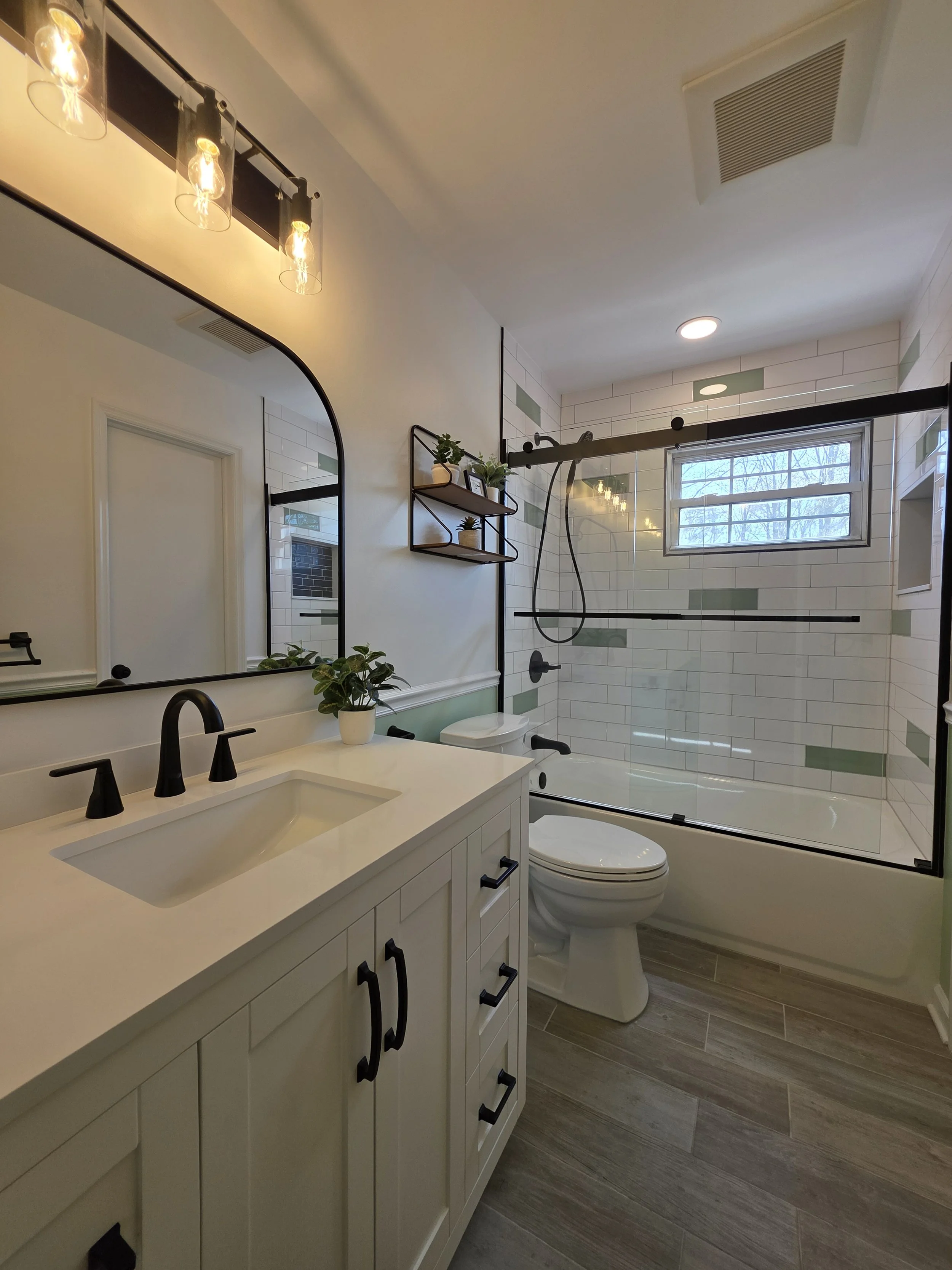 View along the white vanity toward the tiled bathtub, glass enclosure and high window.