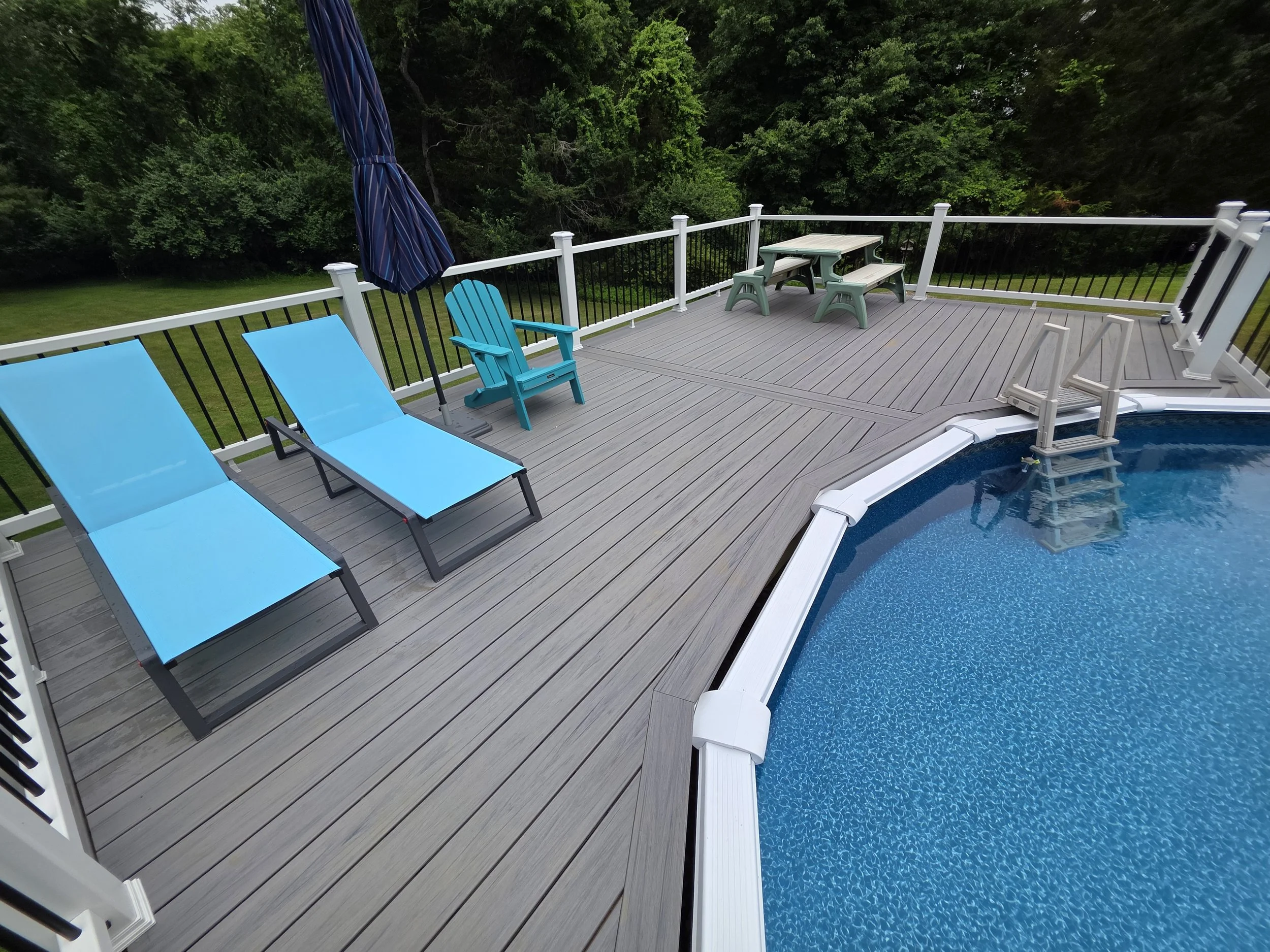Wide view of Lorraine and Ray’s gray pool deck beside a round above-ground pool, with white railings, dark balusters and a seating area.