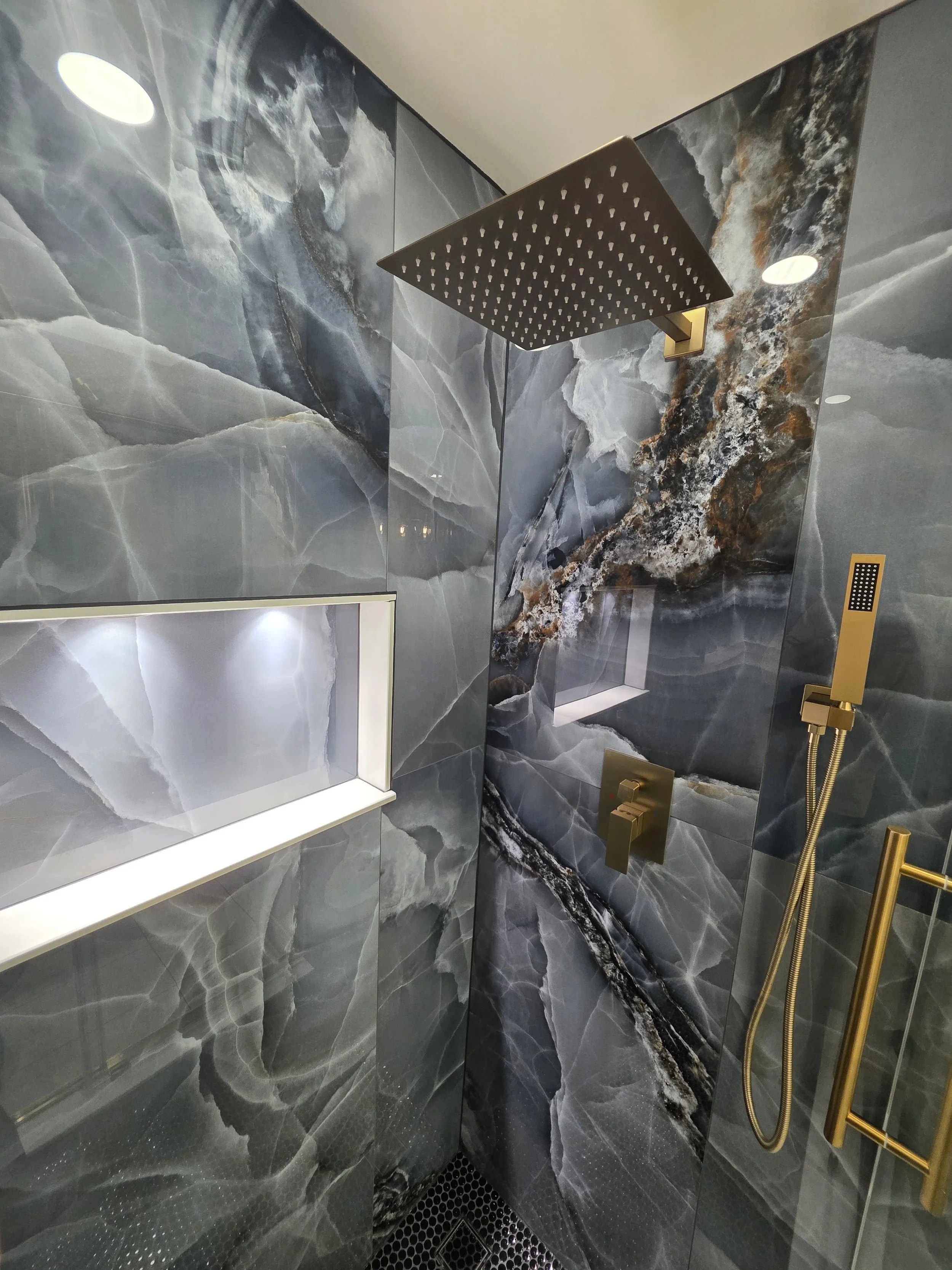 Close view of the gold-tone rain showerhead and handheld spray beside the illuminated shower niche.