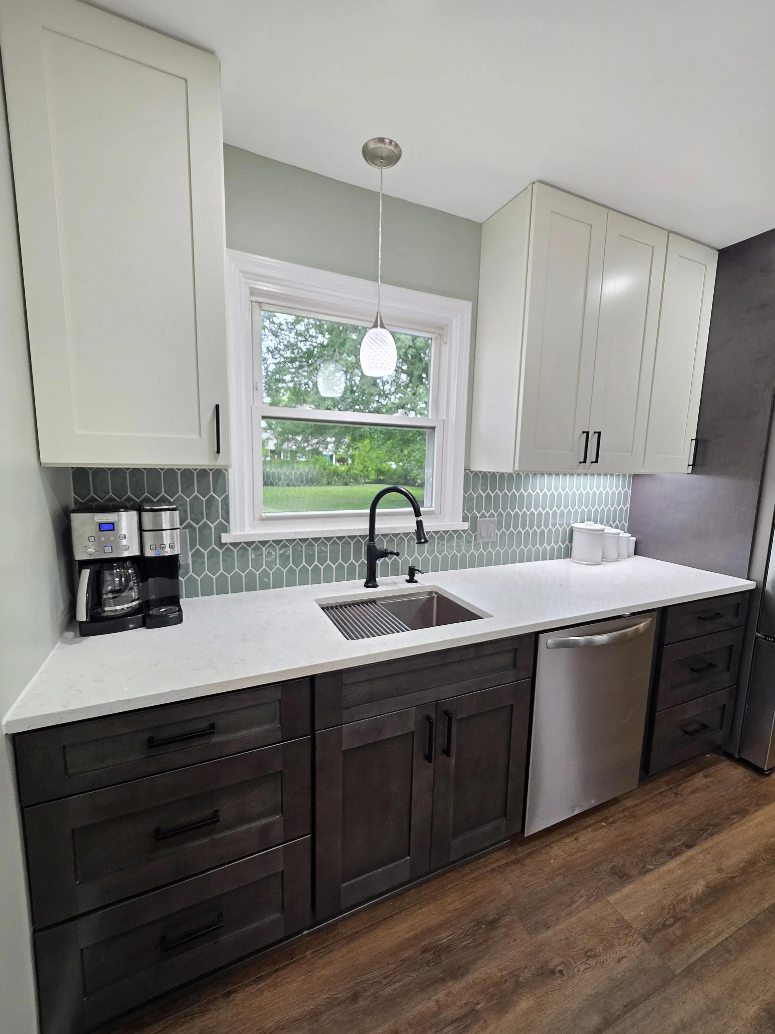 Sink beneath the window, surrounded by light cabinets, patterned backsplash and dark drawers.