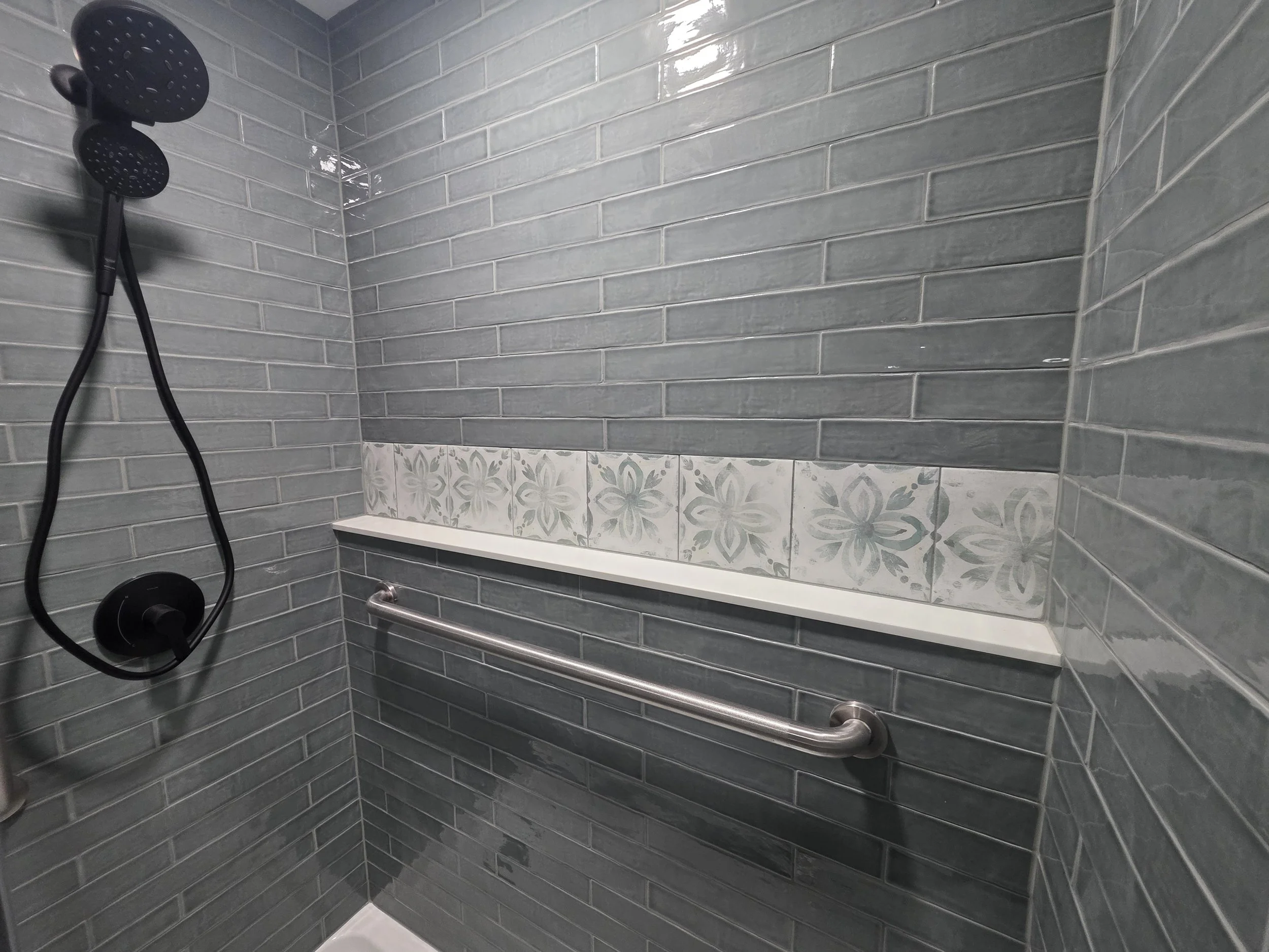 Shower interior with dark fixtures, gray wall tile, patterned ledge and grab bar.