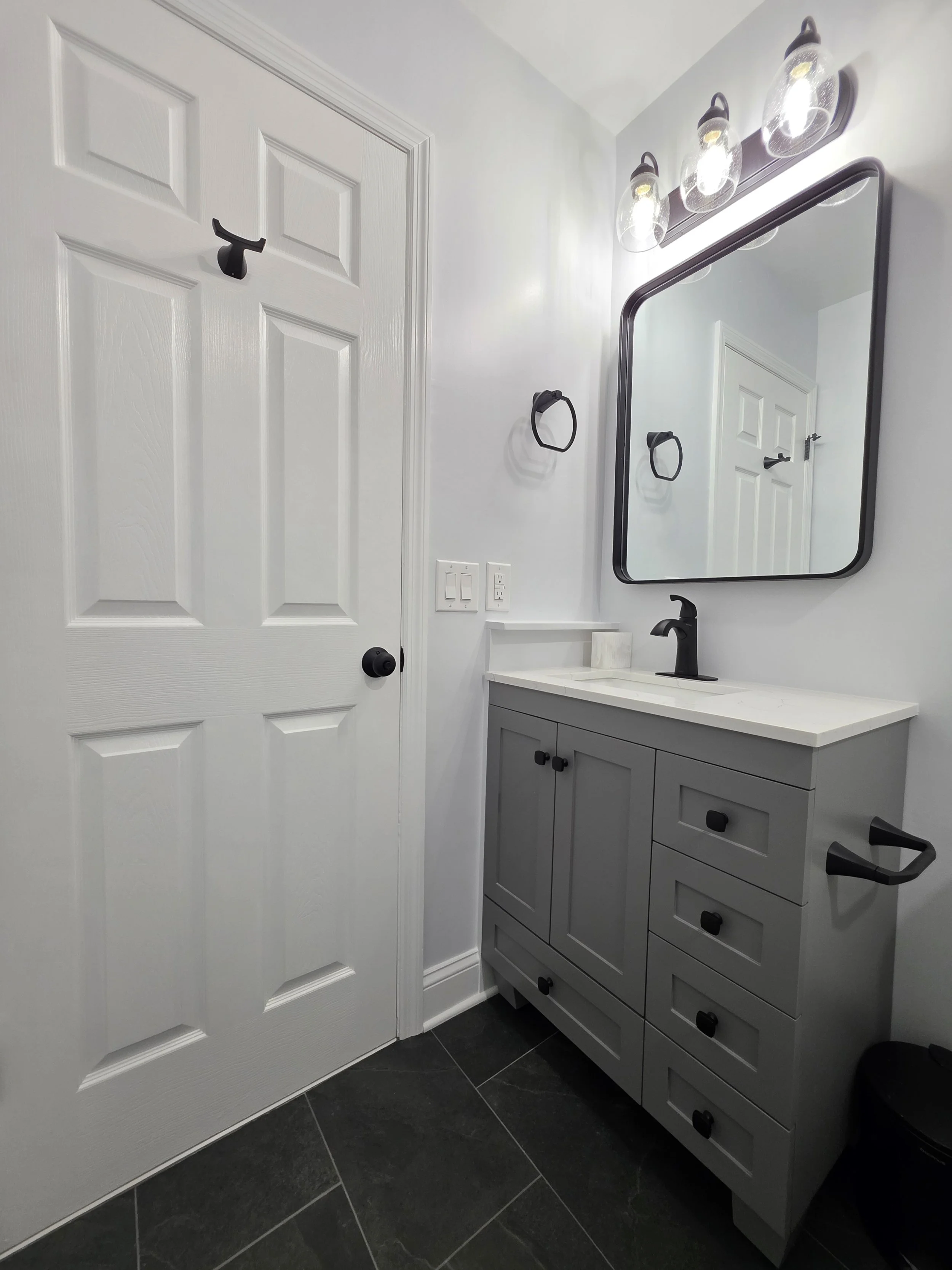 Gray vanity beside the white door, with black faucet, framed mirror and overhead lighting.