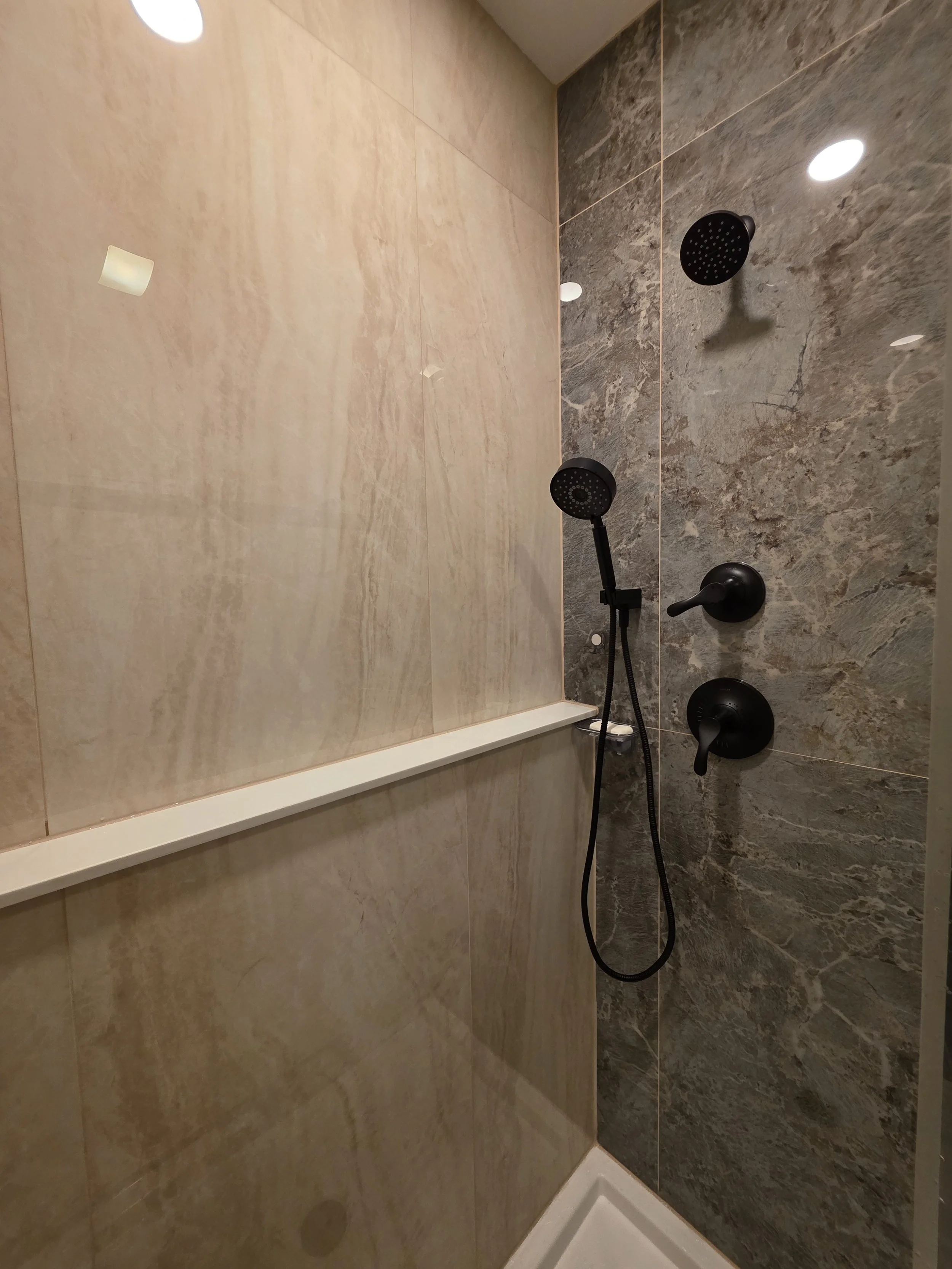 Black showerhead, handheld spray and controls on the textured accent wall beside a long white shower ledge.