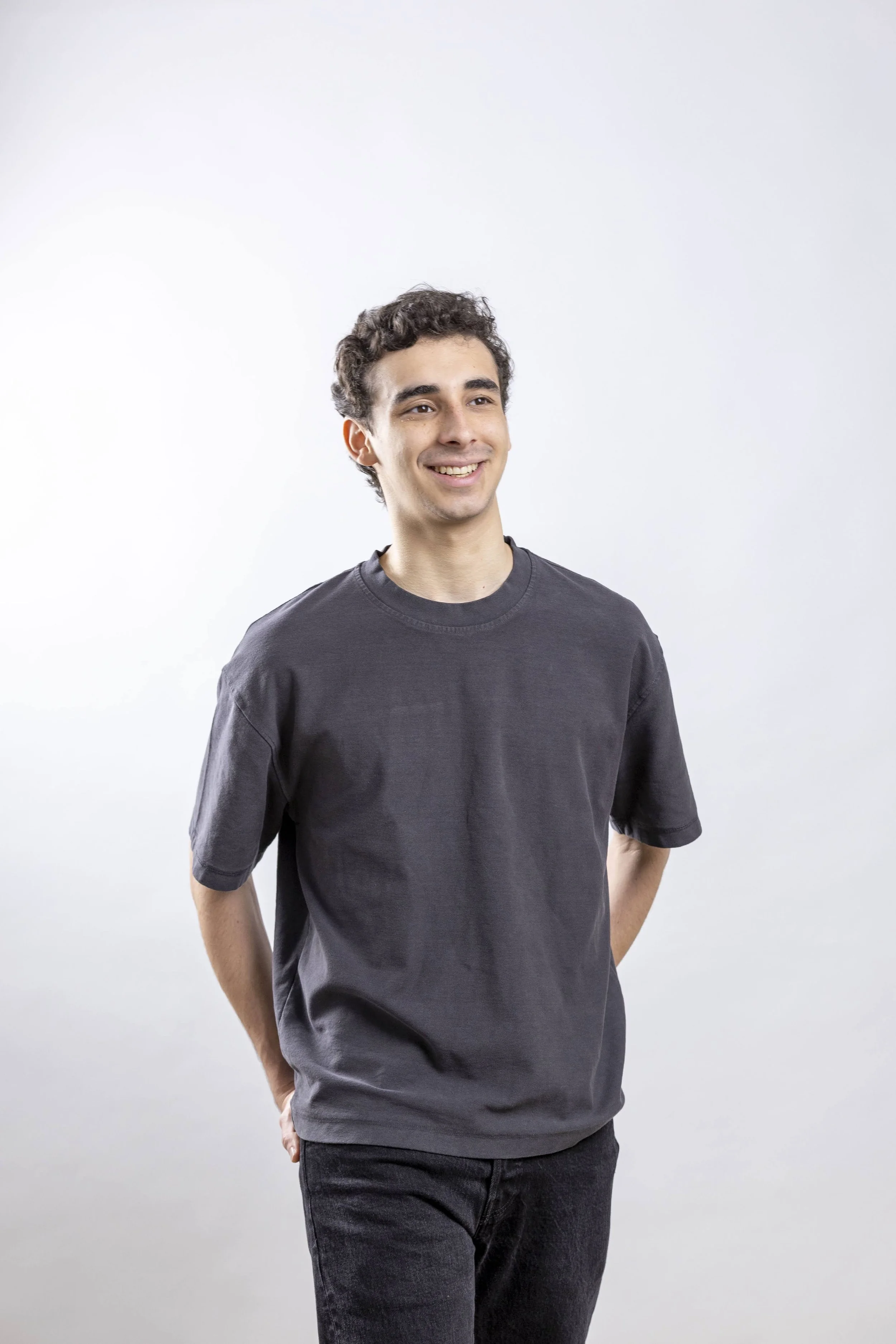 Young man with dark curly hair smiling, wearing a black t-shirt and black jeans, standing against a plain white background.