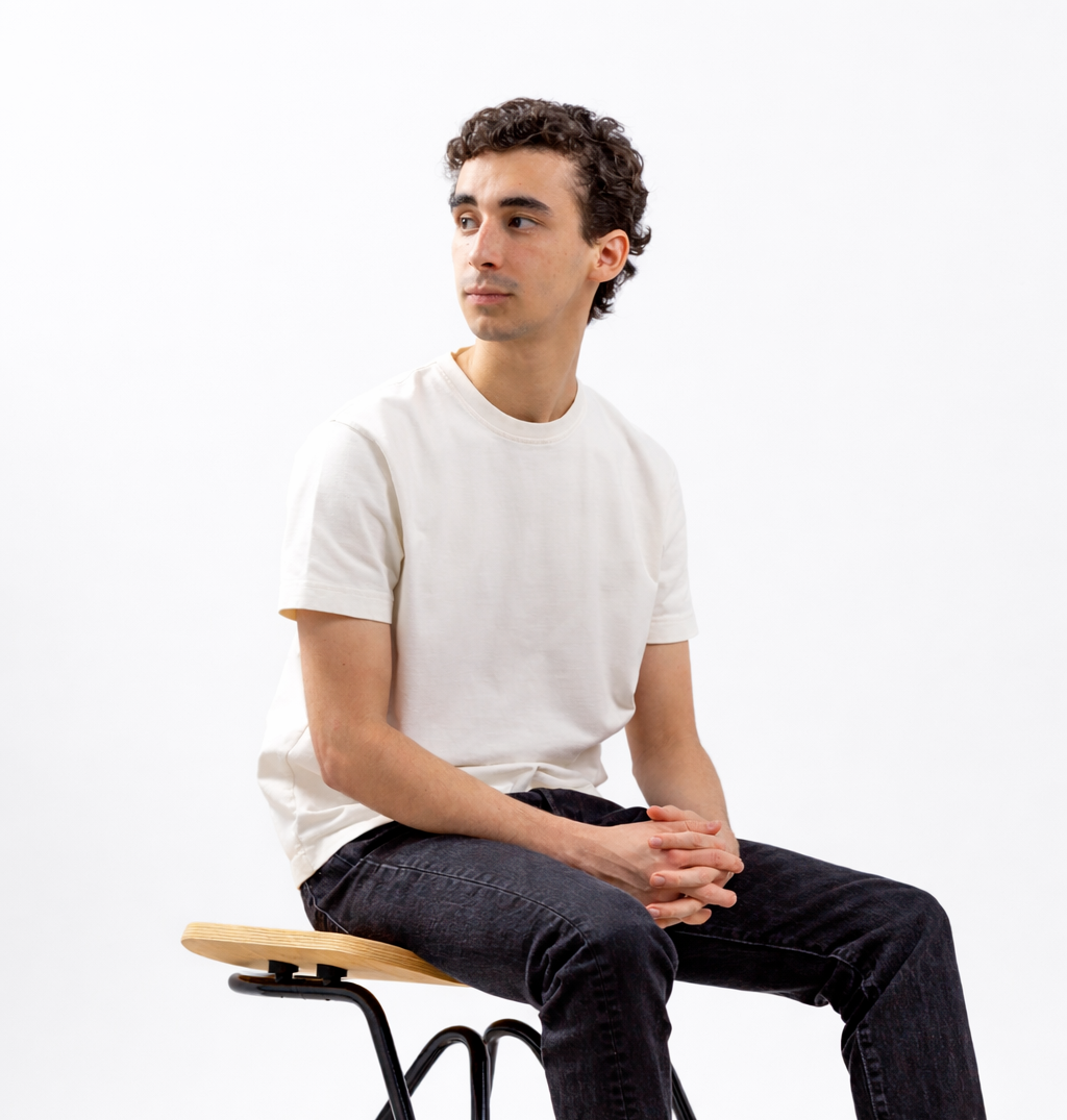 Young man with curly hair sitting on a wooden stool against a white background, wearing a white t-shirt and black jeans, with his hands clasped in his lap, looking to the left.