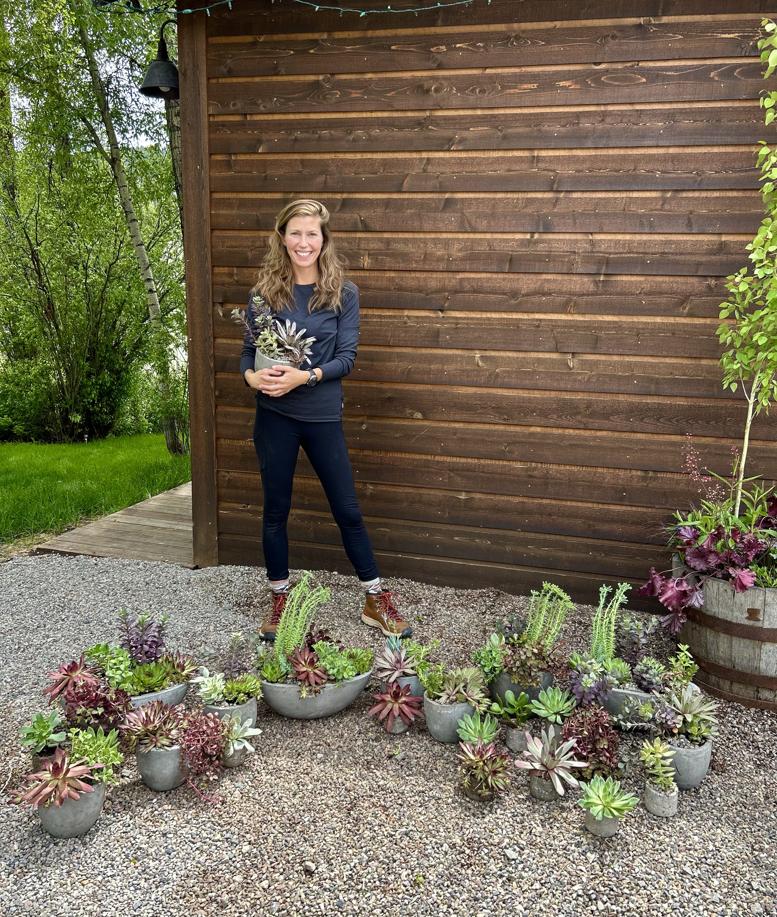 A woman with long hair, wearing a navy sweatshirt and black pants, holding a potted plant, standing outdoors on gravel in front of a wooden wall with various succulent plants in pots at her feet.