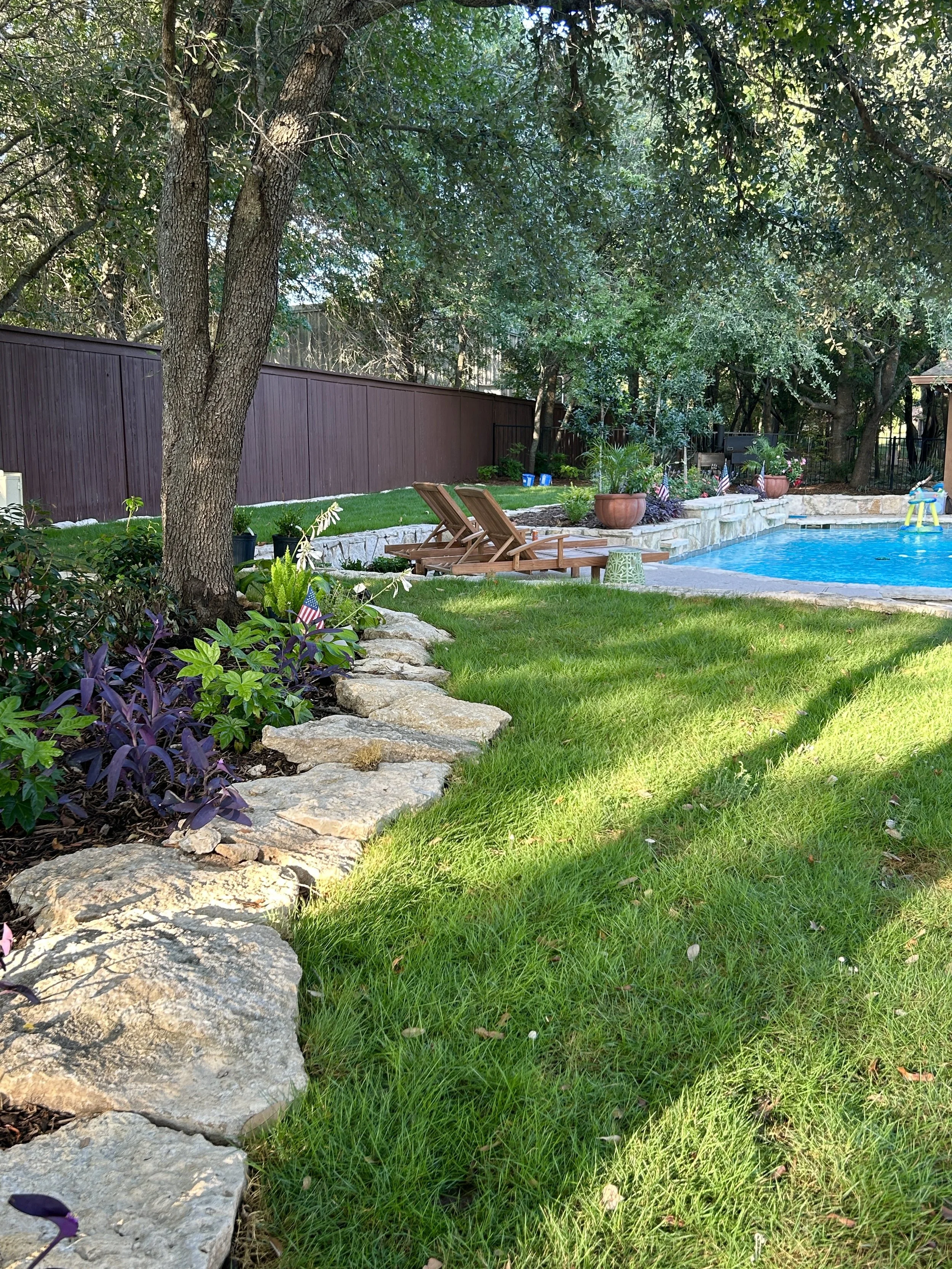 A backyard with a green lawn, large trees, a stone edge garden with purple and green plants, a swimming pool, umbrella, and wooden lounge chairs.