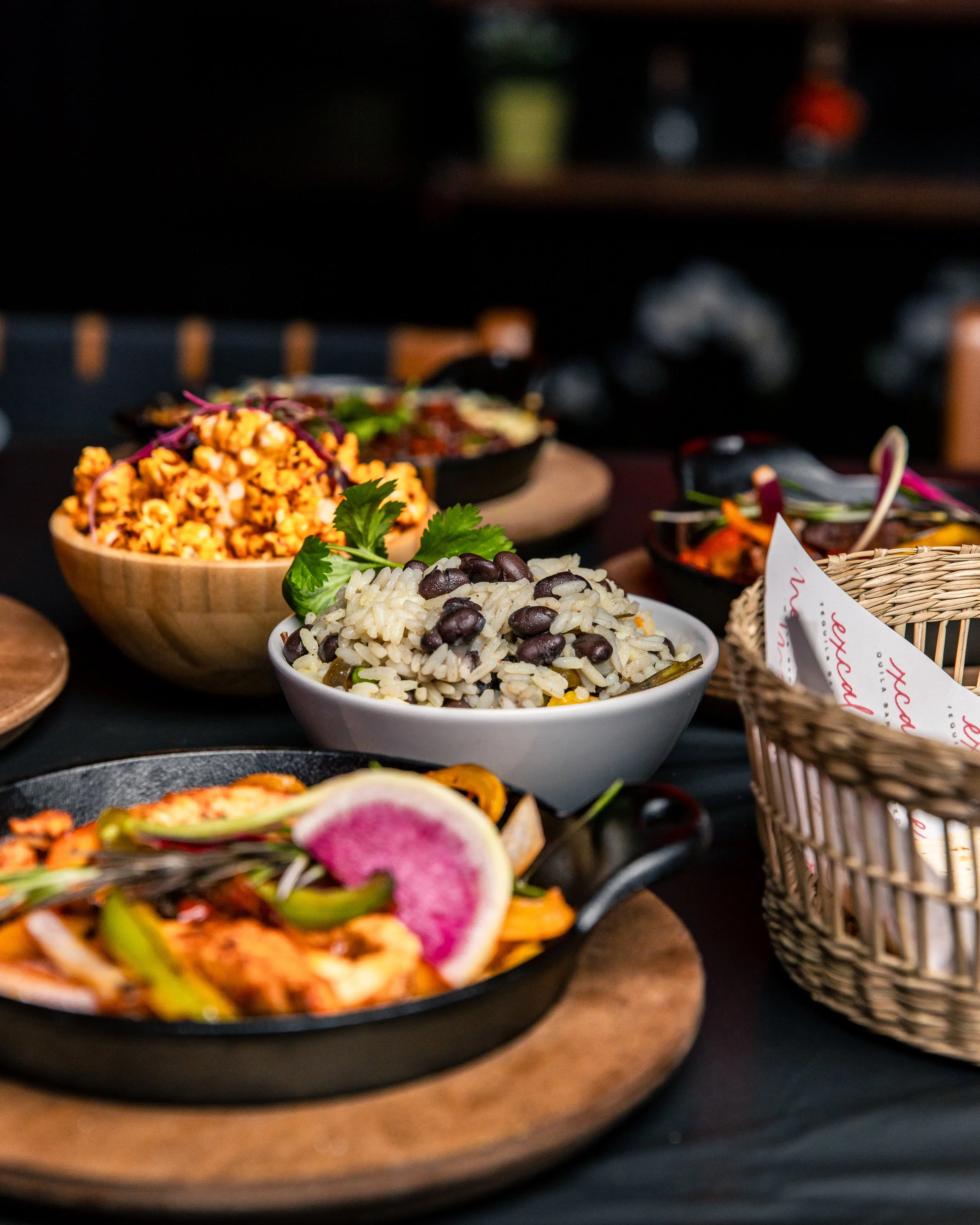 Various dishes of food, including rice, beans, vegetables, and garnishes, presented on plates and bowls, with a dark background.