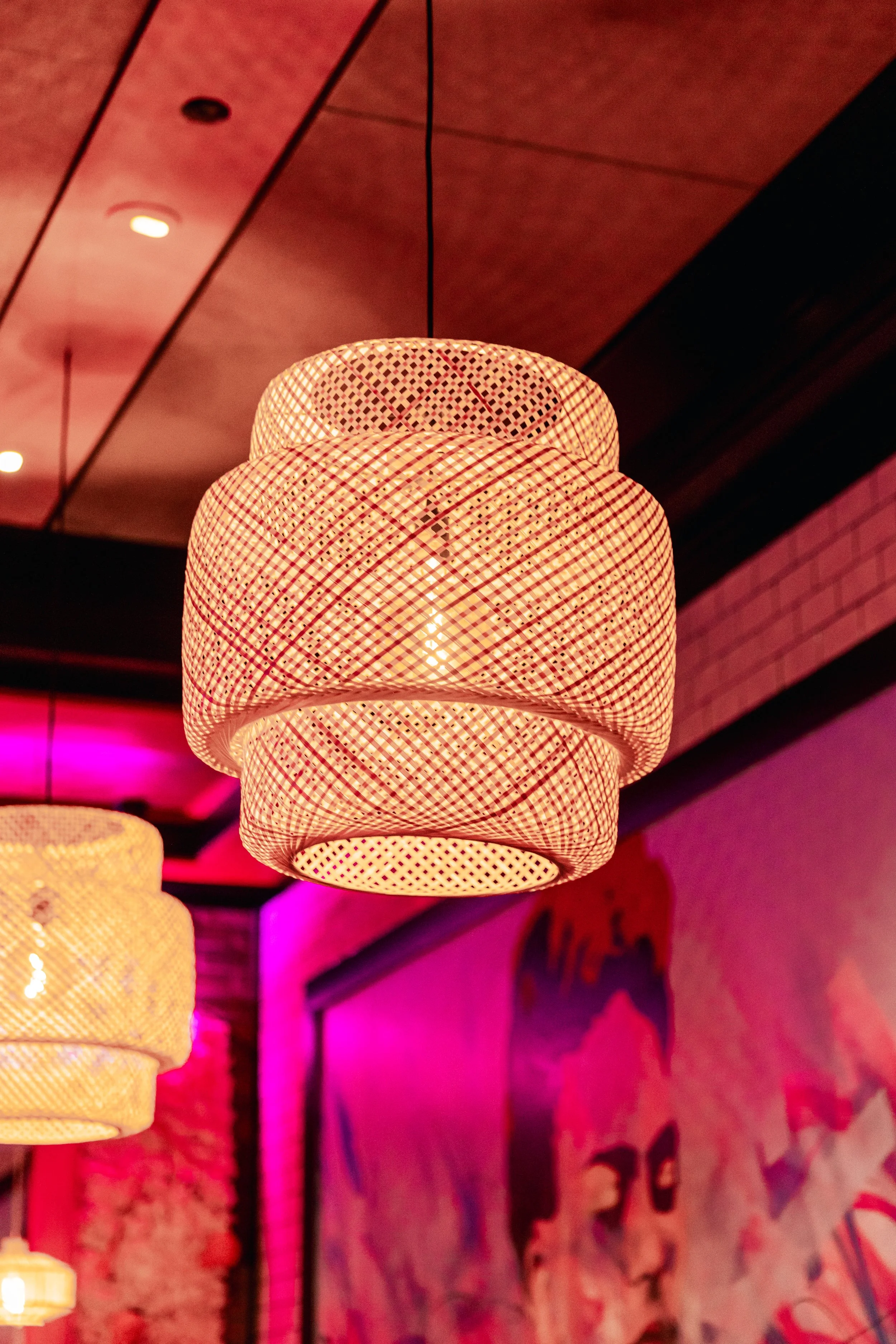 Close-up of a woven, basket-shaped hanging light fixture. The background shows a brick wall and colorful ambient lighting.